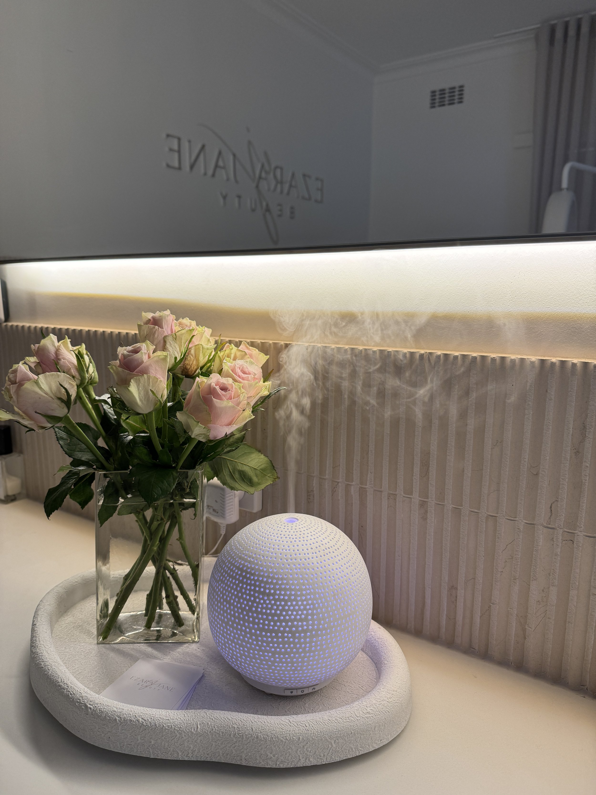 A white spherical humidifier emitting mist, placed on a white tray with a glass vase of pink and white roses on a bedside table.