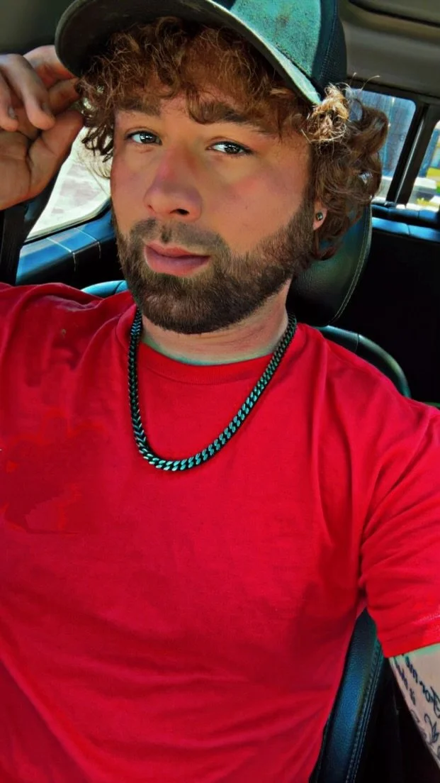 A young man with curly hair and a beard, wearing a red T-shirt, black and turquoise chain necklace, and a baseball cap, sitting inside a vehicle and looking at the camera.