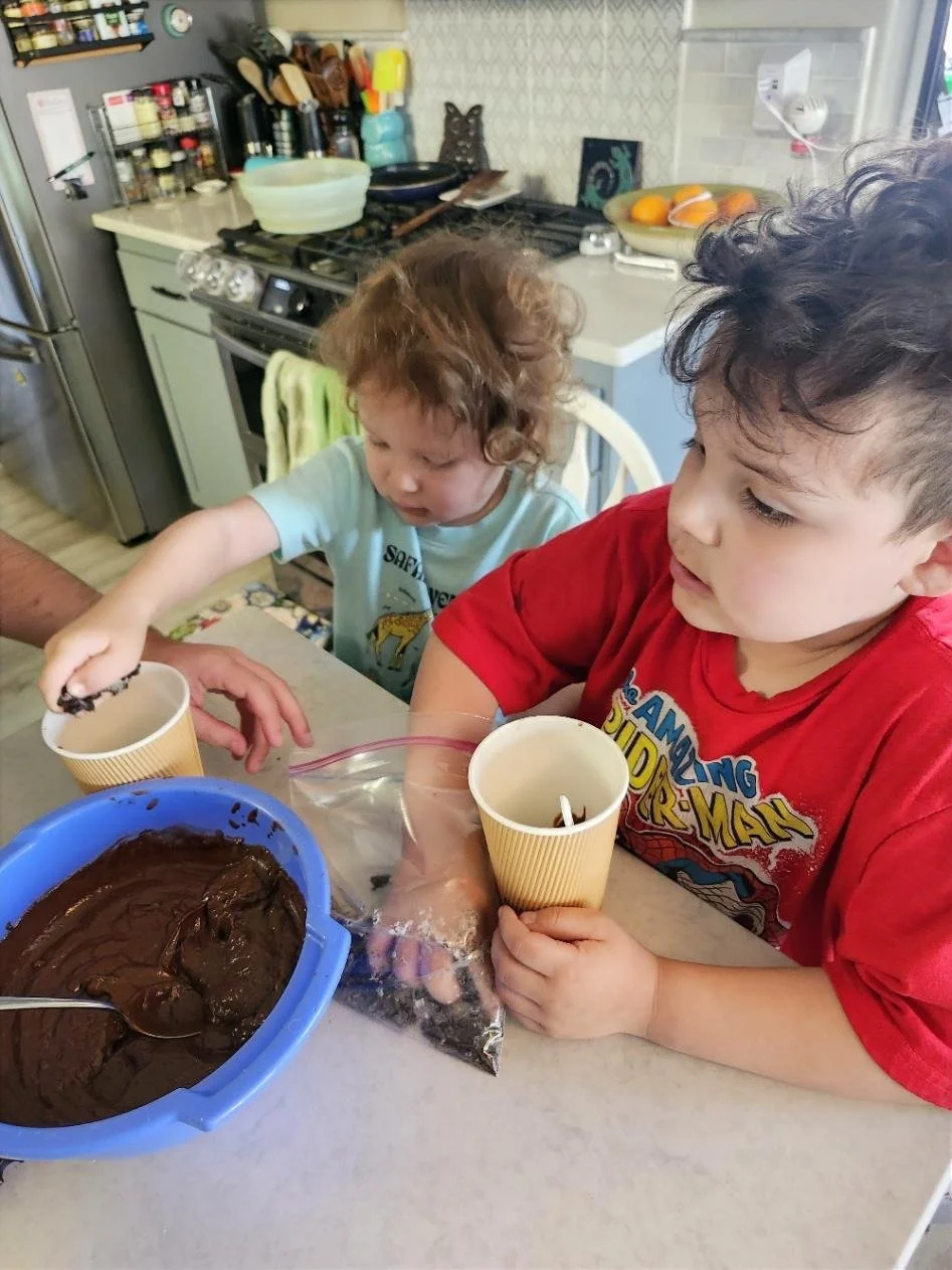 Child stirring dough in a blue mixing bowl while another child stands nearby in a kitchen.