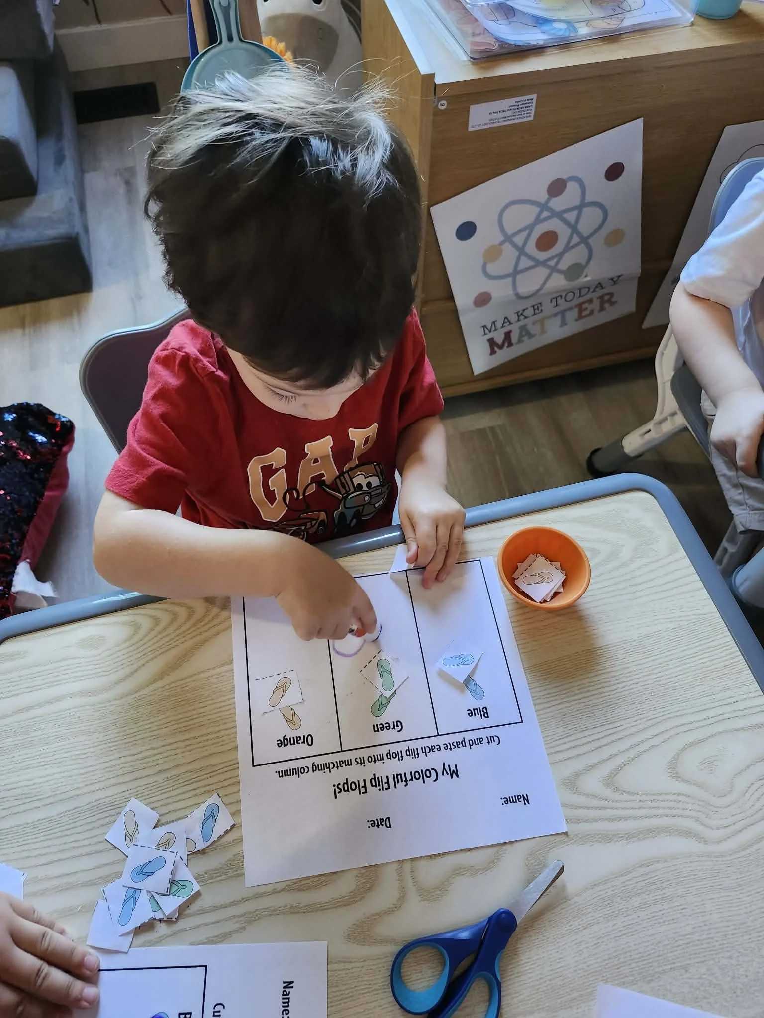 A child with a pink heart covering their face is drawing a poster on a wooden table. The poster has a title reading 'ALL ABOUT' and includes boxes and drawings related to food, with a ruler and marker in the child's hands. There are kitchen items and a paper bag on the table.