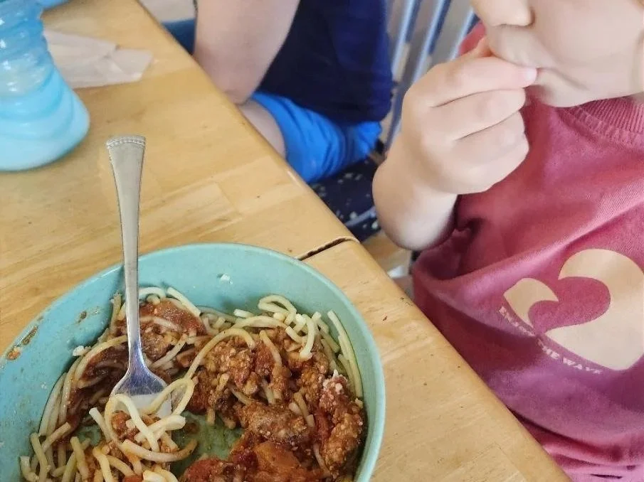 A young child wearing a light gray T-shirt sits at a wooden table with a blue plate containing partially eaten pieces of chicken, grapes, and a small piece of cake. A fork rests on the plate, and the child's face is obscured with a heart emoji.
