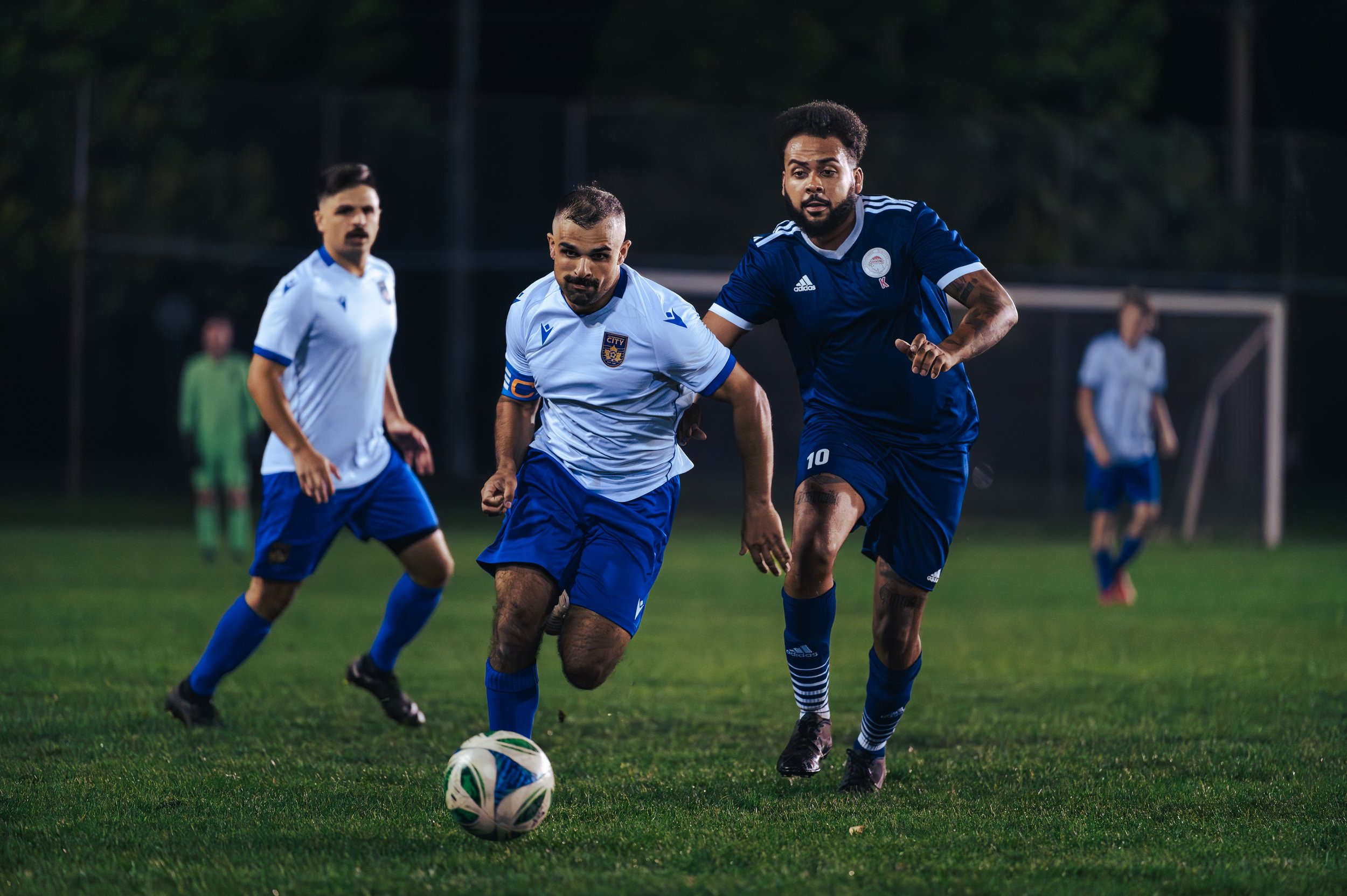 Peterborough City Soccer players chasing a ball during a match on a grassy field at night, with one player in a light blue jersey and another in a dark blue jersey prominently in the foreground.