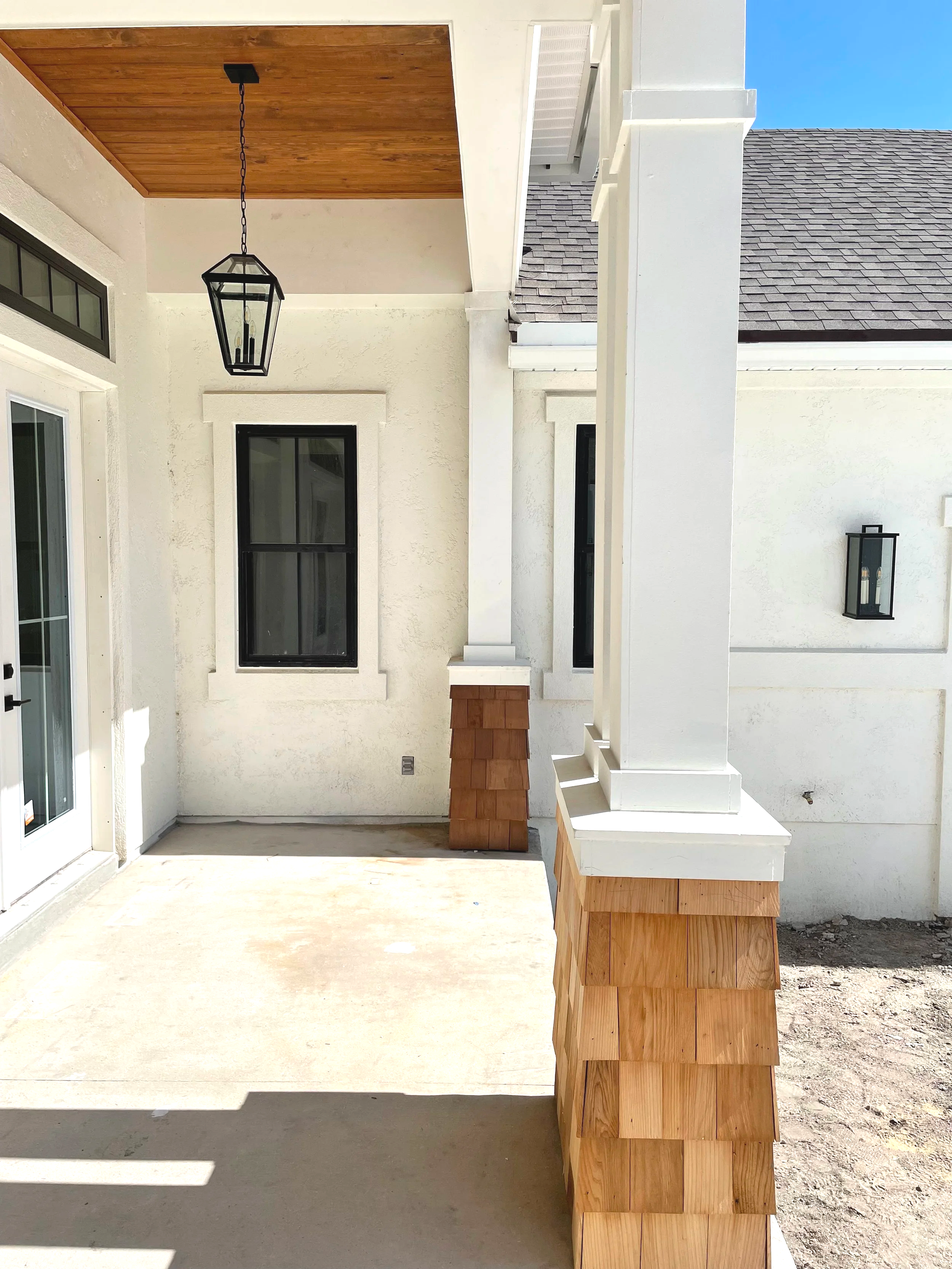 Newly constructed covered porch with white walls, a wood ceiling, a black lantern light fixture, and windows with black frames. There are wooden accents on the porch columns and base, with an unfinished concrete floor.