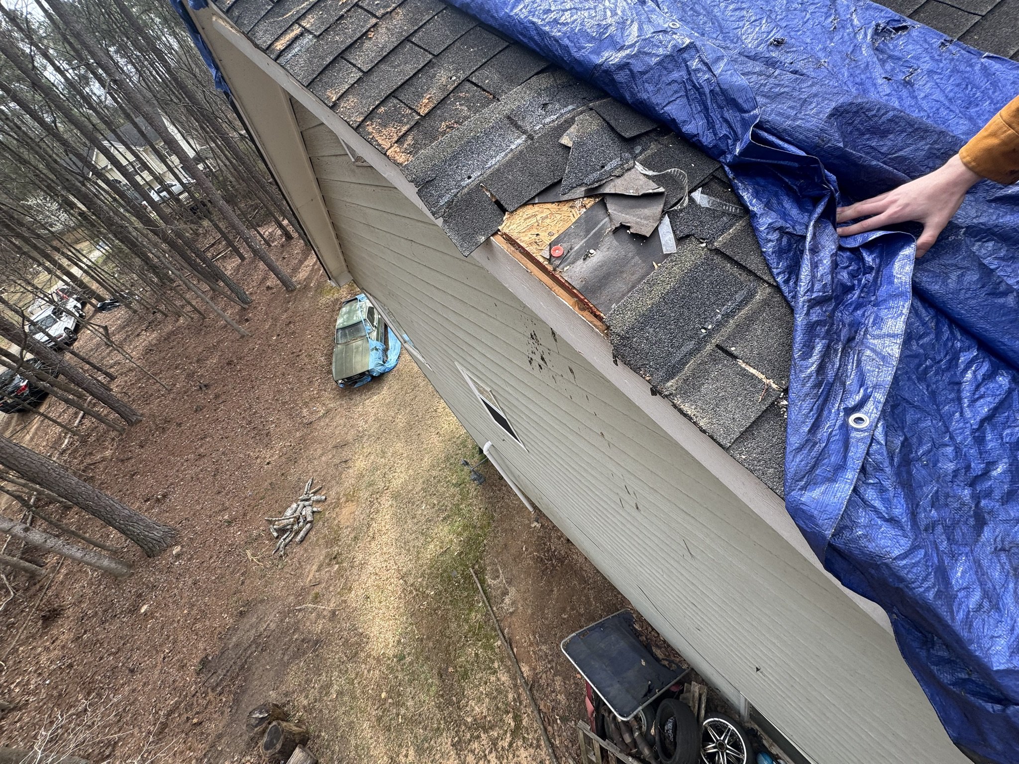A person in a yellow jacket holds a blue tarp over a section of a house roof under repair, with shingles removed and tools on the ground.