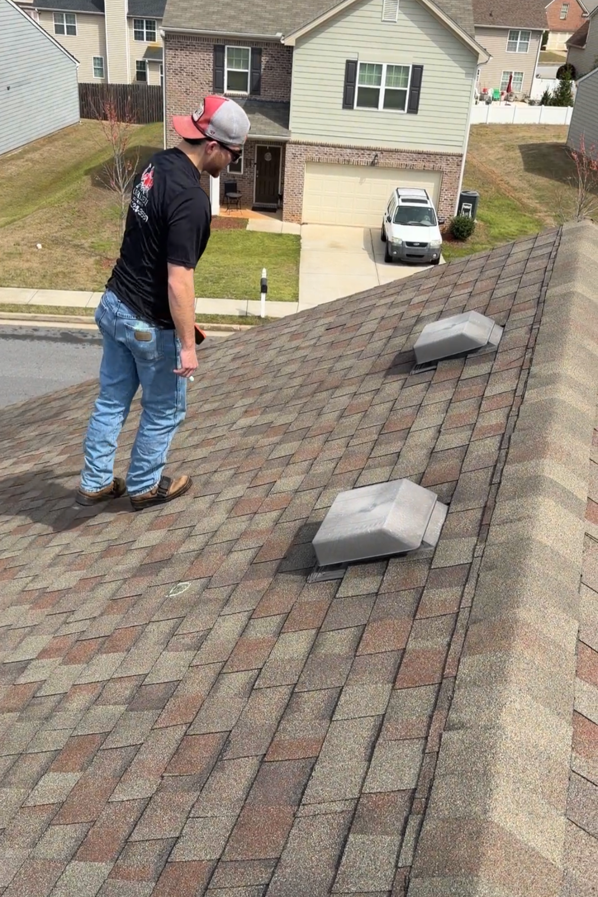 A man in a black jacket, jeans, and a gray cap stands on a residential roof, looking down at the shingles and roof vents, with a suburban neighborhood visible in the background.