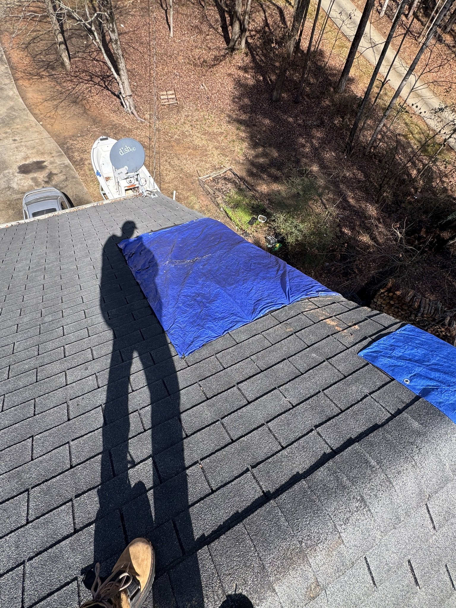 Top-down view of a residential rooftop with a shadow of a person, some blue tarps, a satellite dish labeled "dish tv", a car, and a backyard with trees and a paved driveway.
