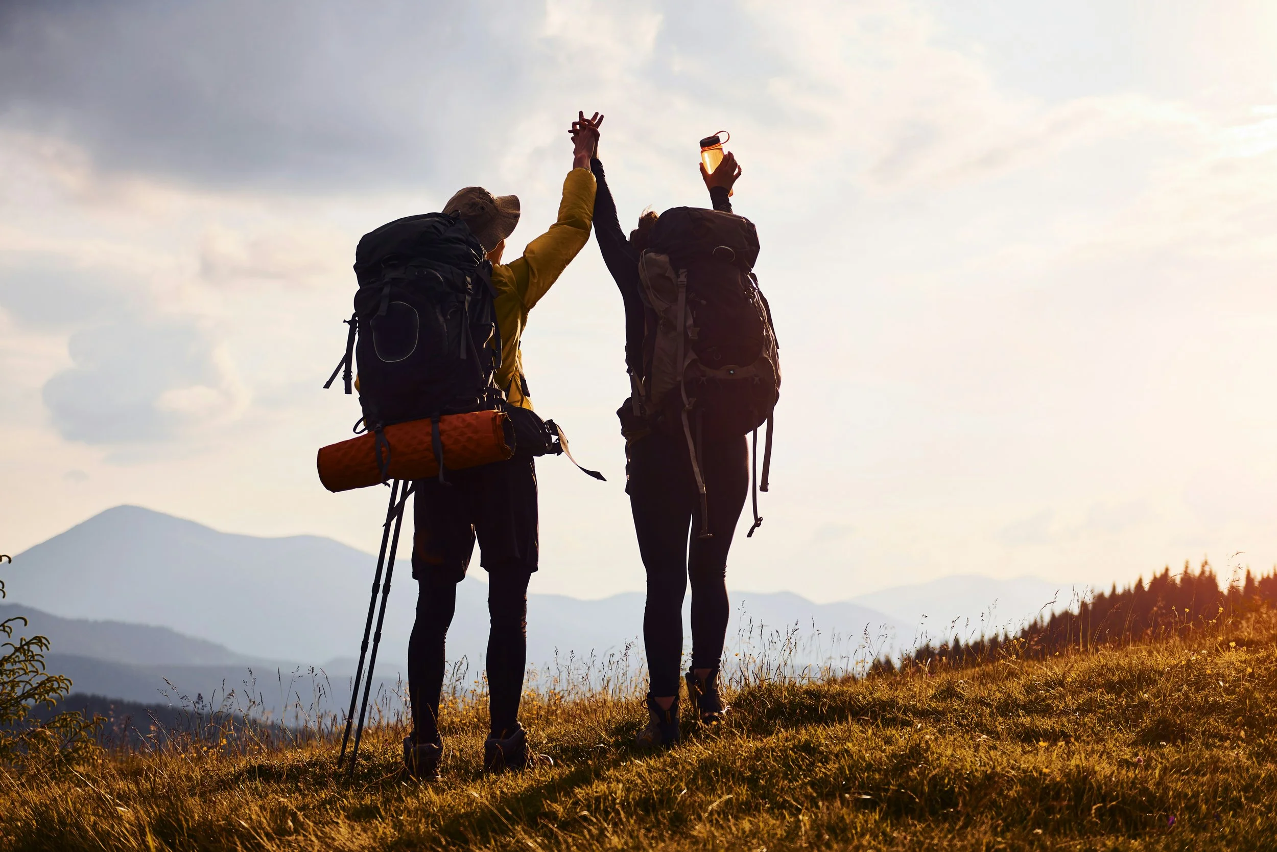 Two hikers with backpacks and trekking poles giving a high five on a mountain trail during sunset.