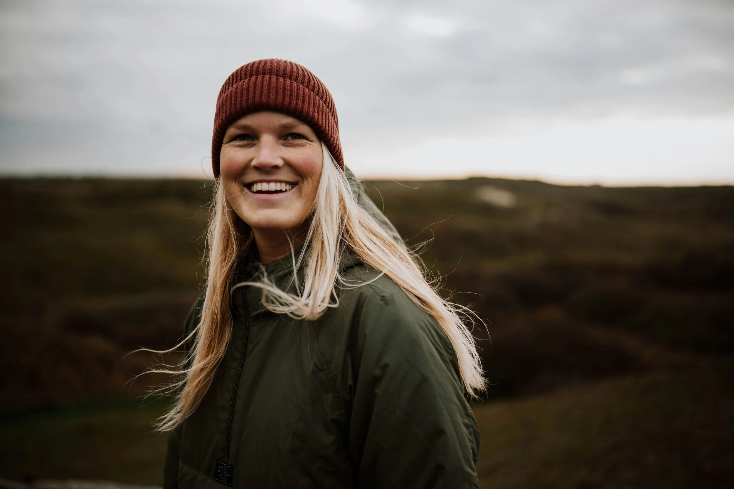 A smiling woman with long blonde hair wearing a red knit beanie and a green jacket outdoors on a cloudy day.