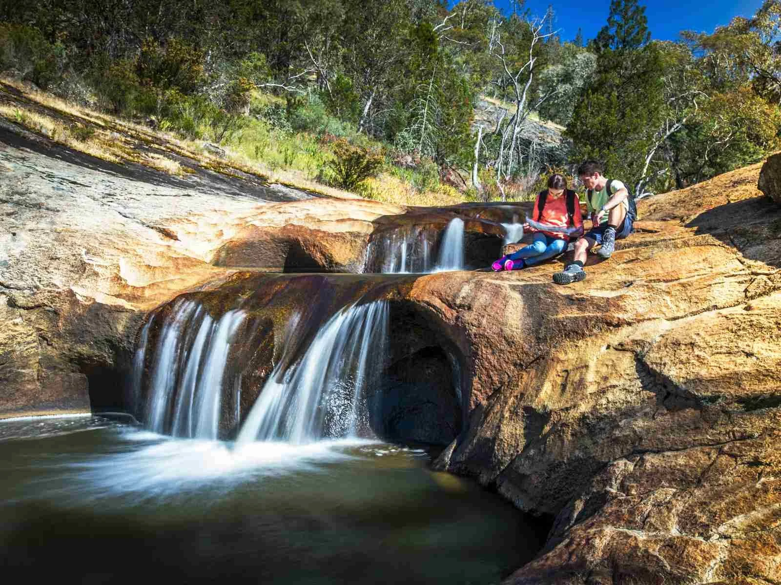 Two children sitting on a rock near a small waterfall in a forested area, looking at a map or book.
