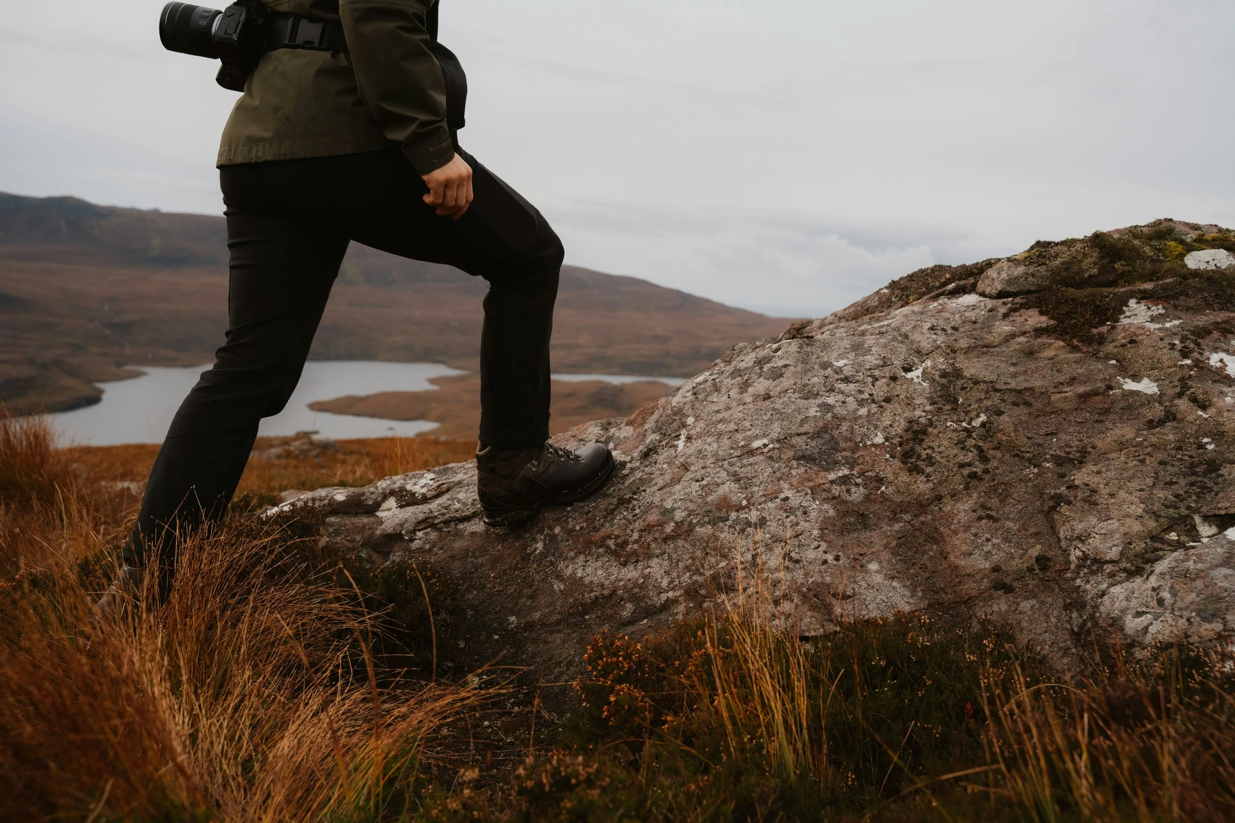 A person standing on a rocky terrain with a lake and mountains in the background under a cloudy sky.