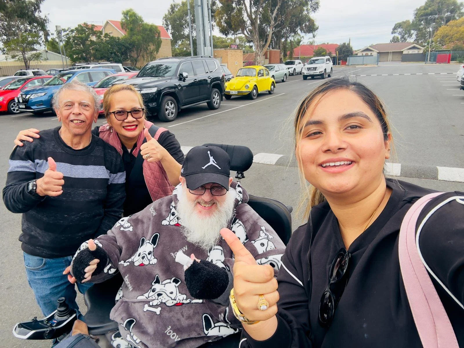 Group photo of four people, including a man with a beard and a woman with glasses giving thumbs up, taking a selfie in a parking lot with various parked cars and a yellow vintage car in the background.