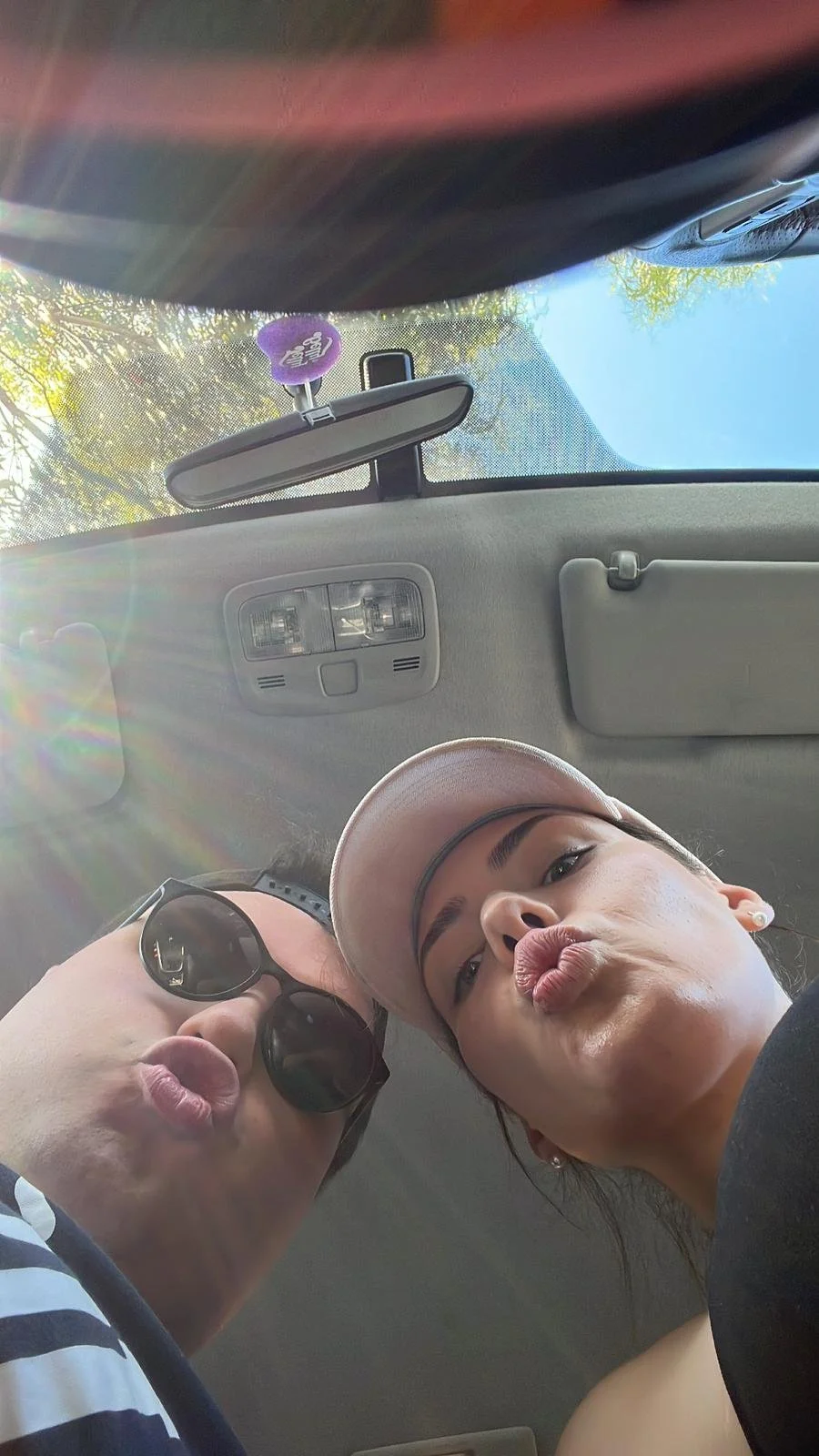 Two women making duck faces taking a selfie inside a car. One woman is wearing sunglasses and a striped shirt, the other is wearing a pink cap and black top.