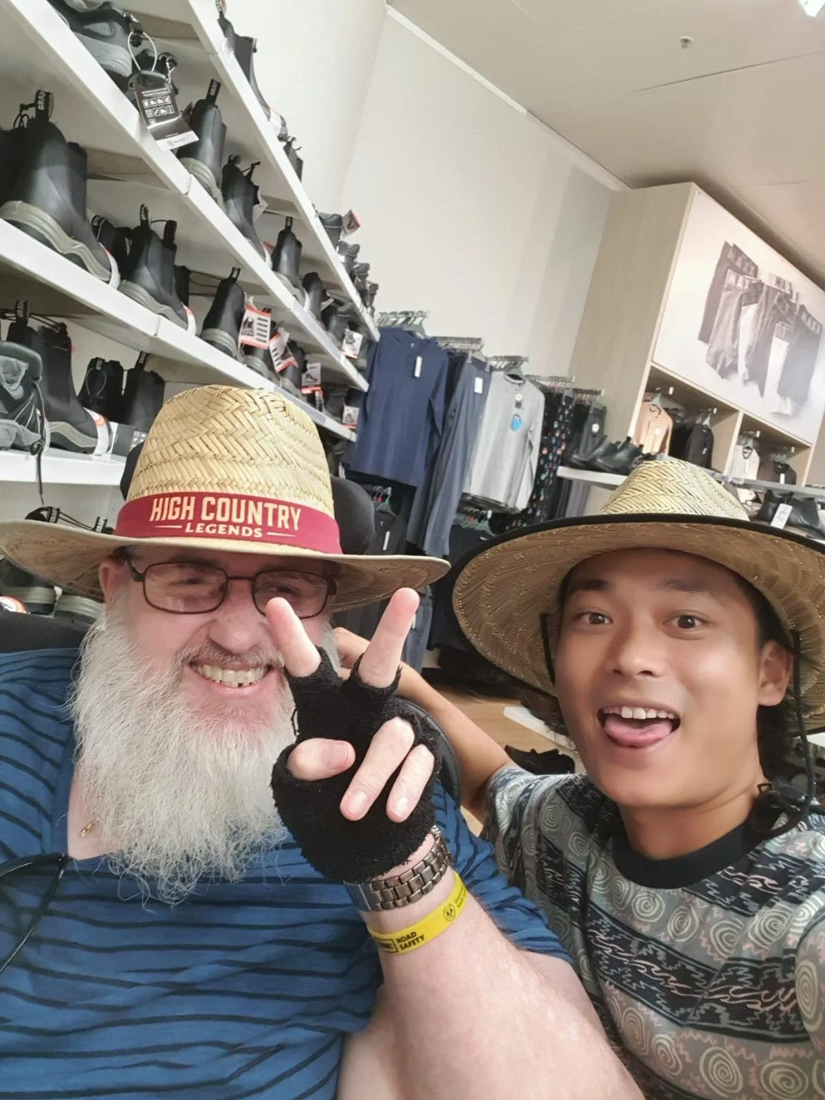 Two smiling men wearing straw hats, with one making a peace sign and the other sticking out his tongue, in an outdoor store or market with shoes and clothing on the shelves behind them.