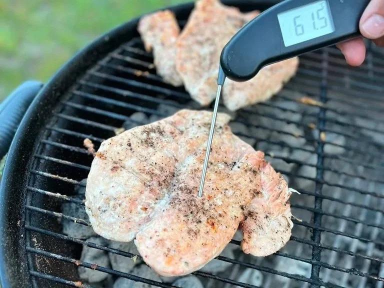 A hand holding a digital meat thermometer over a piece of seasoned chicken on a grill, with other chicken pieces cooking in the background.