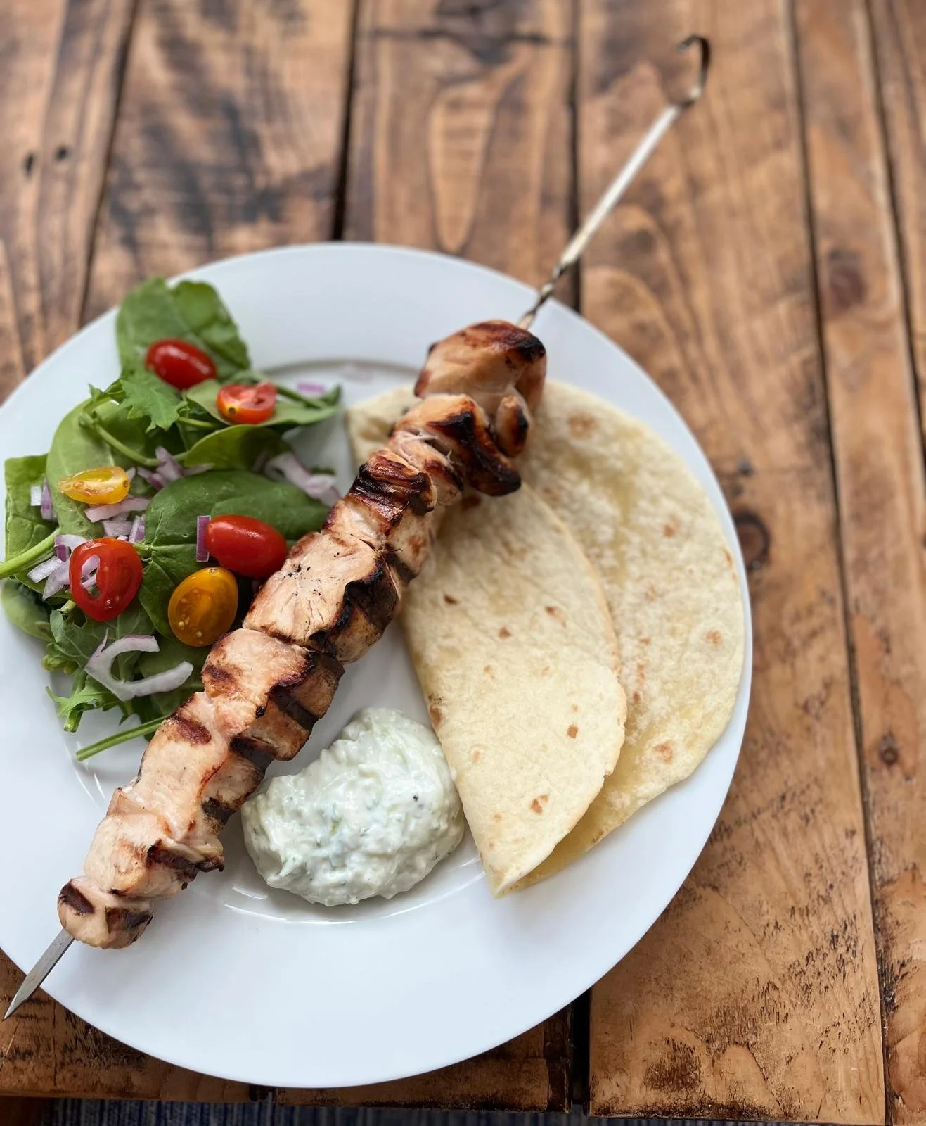 A white plate of grilled chicken kebab skewers, chopped salad with cherry tomatoes and greens, two pita bread pieces and a side of tzatziki sauce, all arranged on a wooden table.