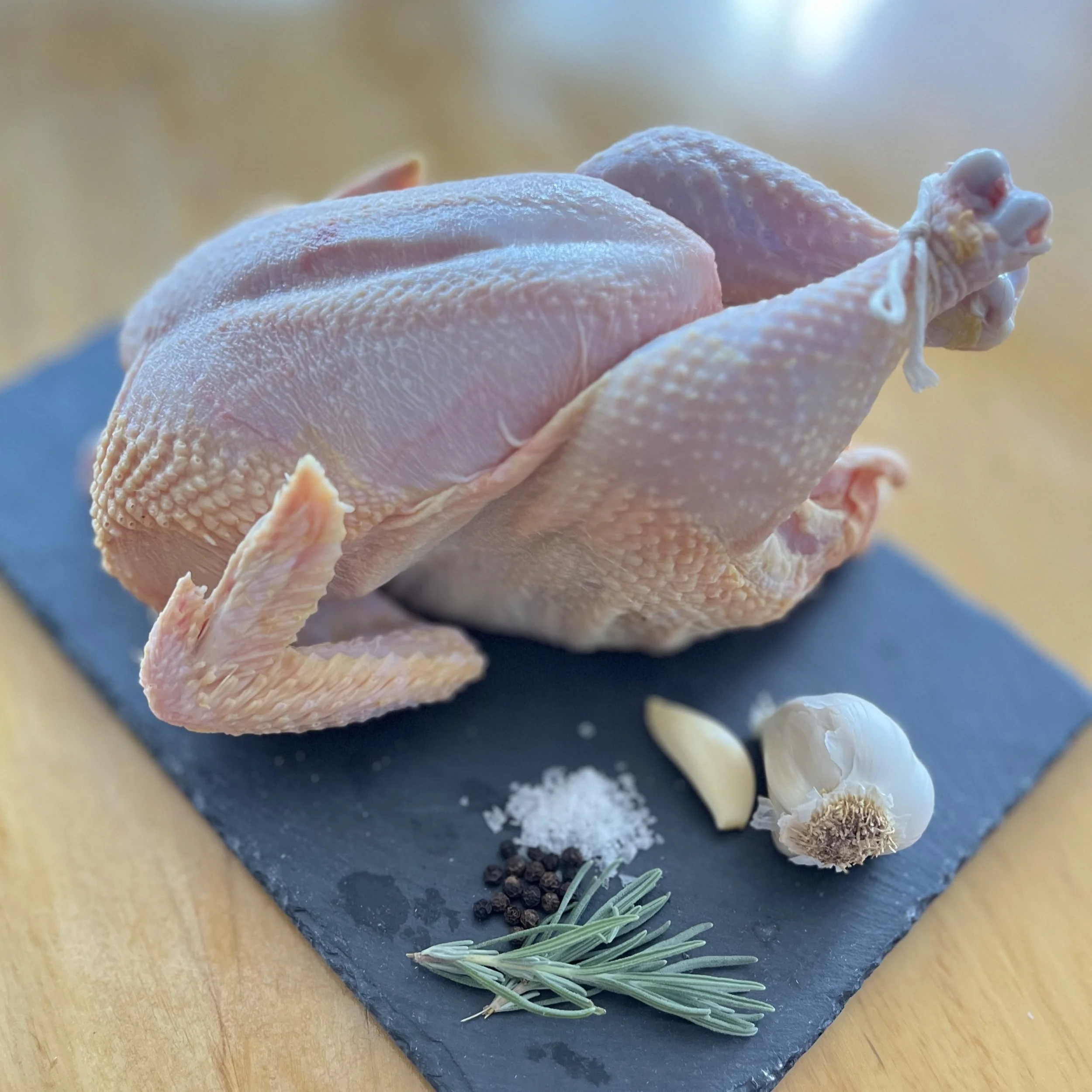 Raw whole chicken with garlic, salt, pepper, rosemary, and garlic cloves on a black slate board.