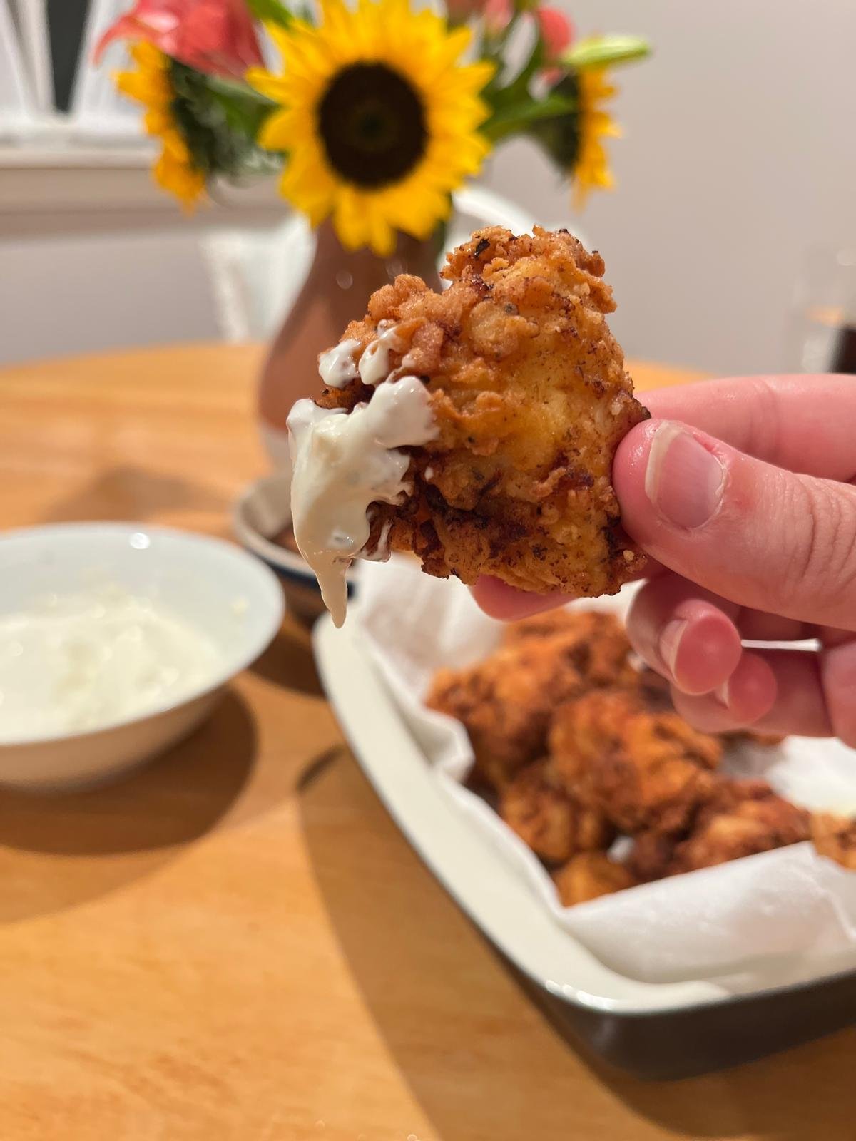 A hand holding a piece of fried chicken with white ranch on it. In the background, there is a plate of fried chicken, a bowl of ranch dip, and a vase with sunflowers on a wooden table.