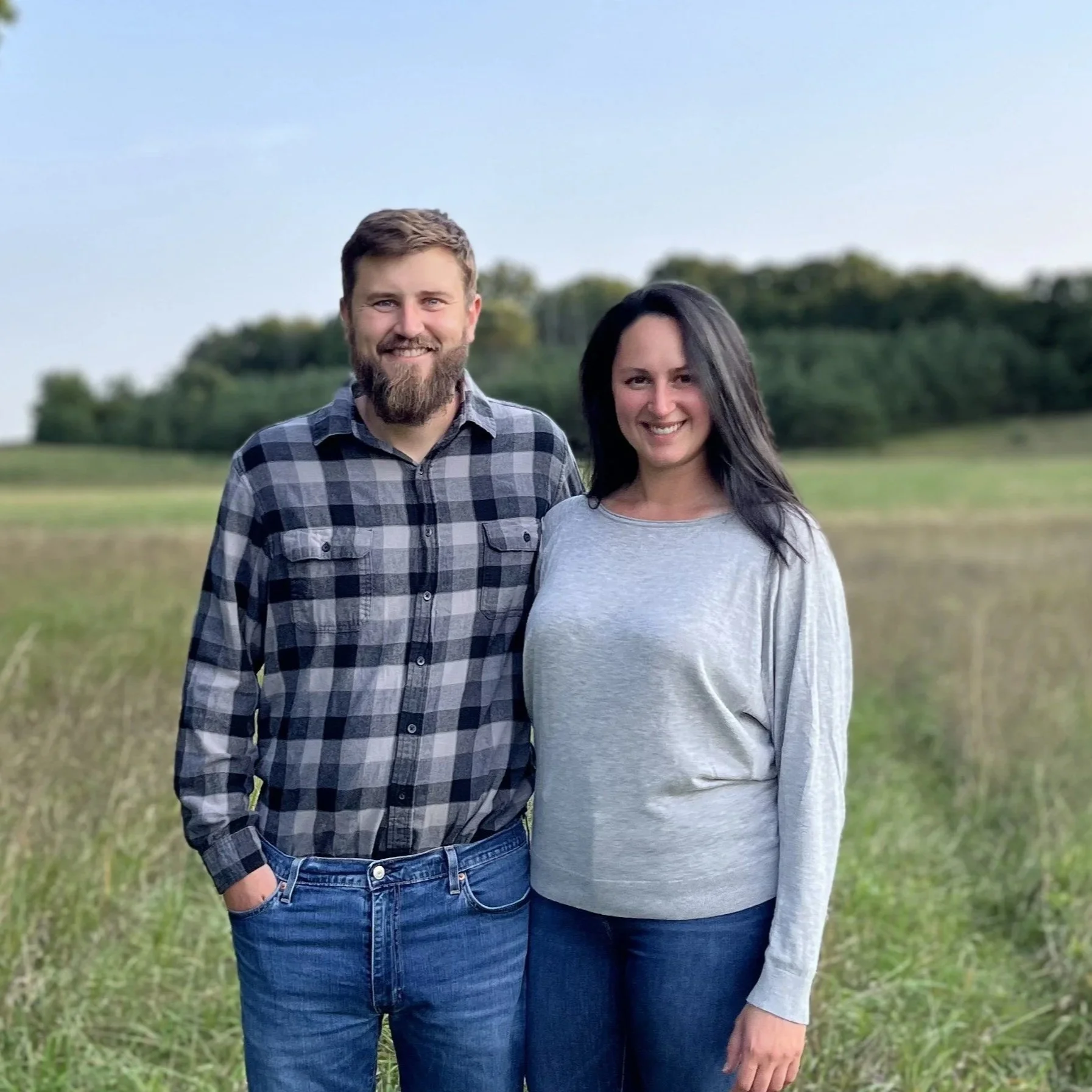 A man and woman standing outdoors in a grassy area with trees in the background, smiling at the camera.