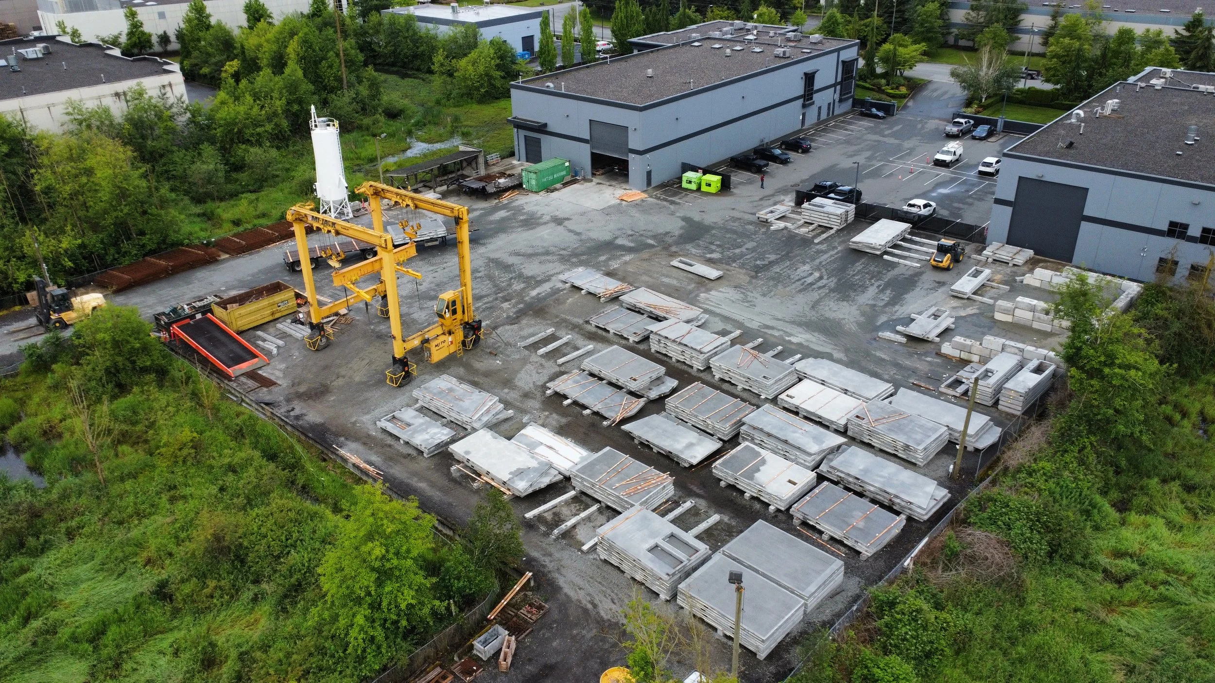An aerial view of the Baseline Concrete warehouse yard with concrete slabs, a yellow crane, and parked cars next to industrial buildings surrounded by green trees.