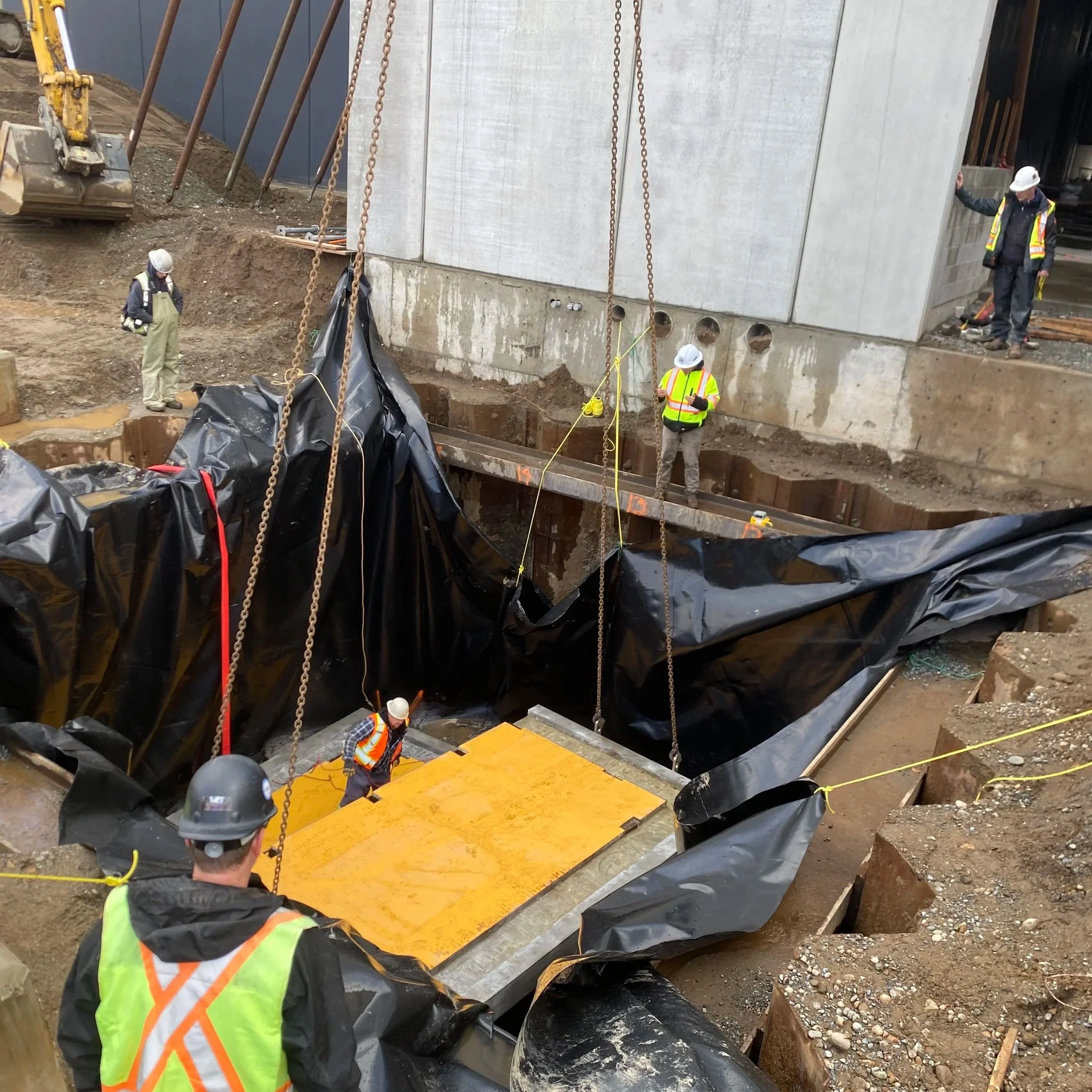 Construction workers wearing safety vests and helmets working on a construction site. Some workers are inspecting a large yellow and black mat placed over a construction pit, with black plastic covering the area and chains hanging down nearby. A dirt trench and construction equipment are visible in the background.