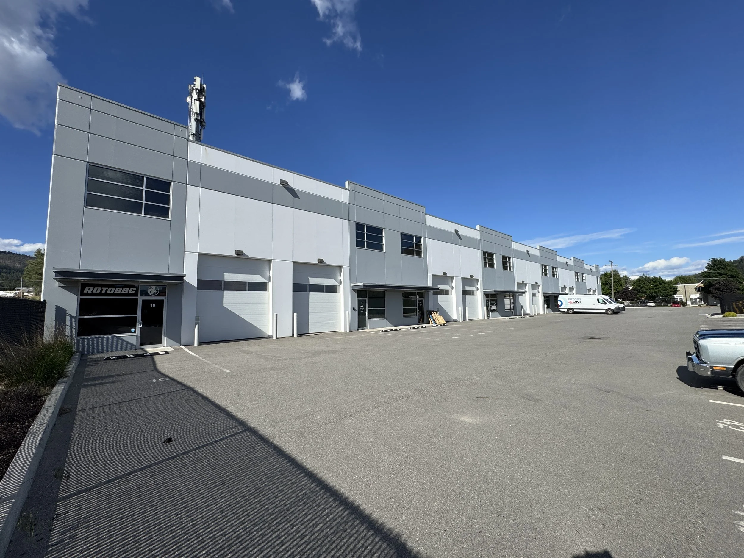 Modern industrial building with gray and white exterior, large windows, and a row of closed garage doors in a parking lot. Clear blue sky with some clouds and a few parked vehicles nearby. The building is made of pre-cast concrete.