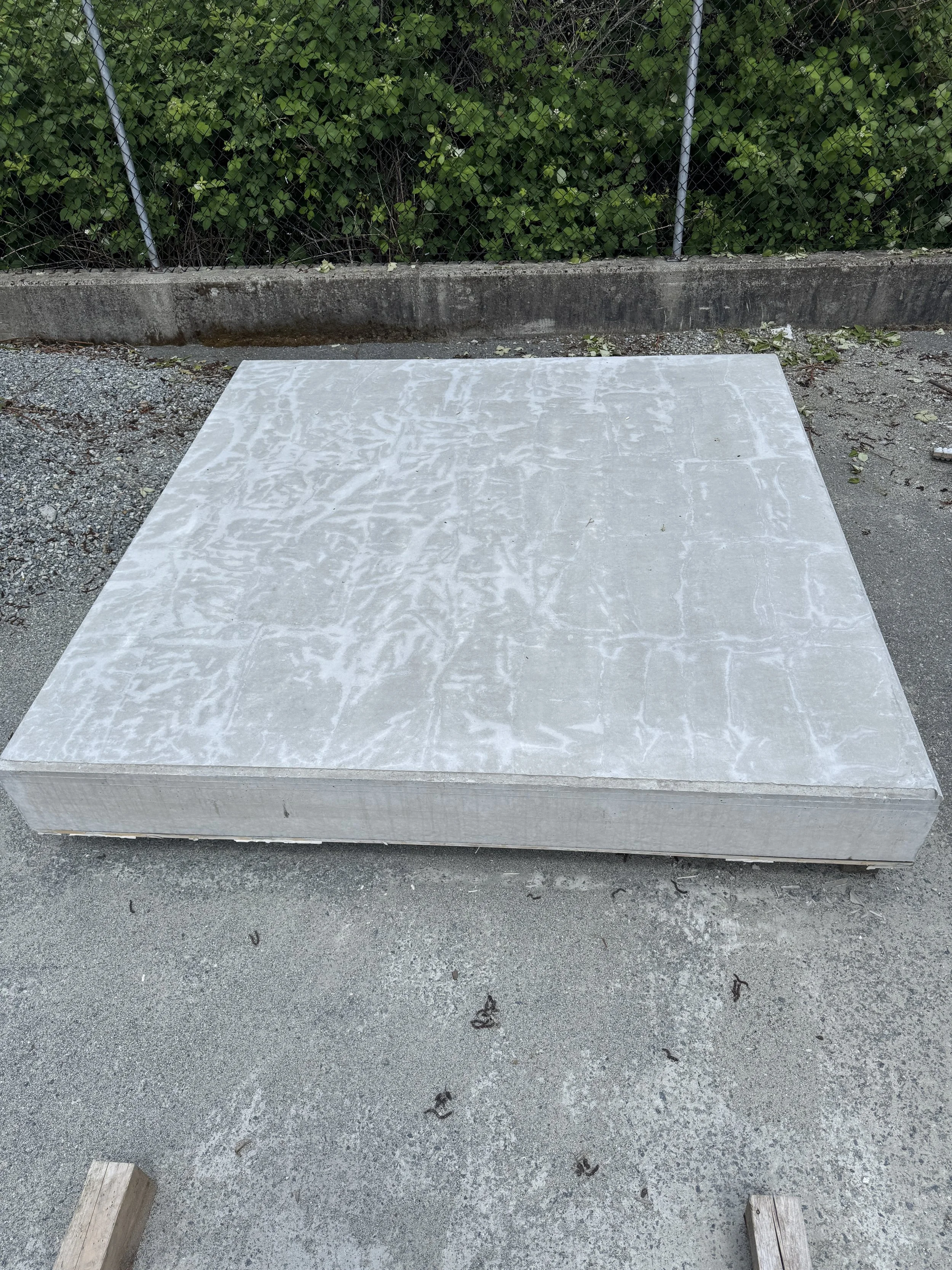 Large rectangular precast concrete wall panel laying on the ground with green foliage and a chain-link fence in the background.