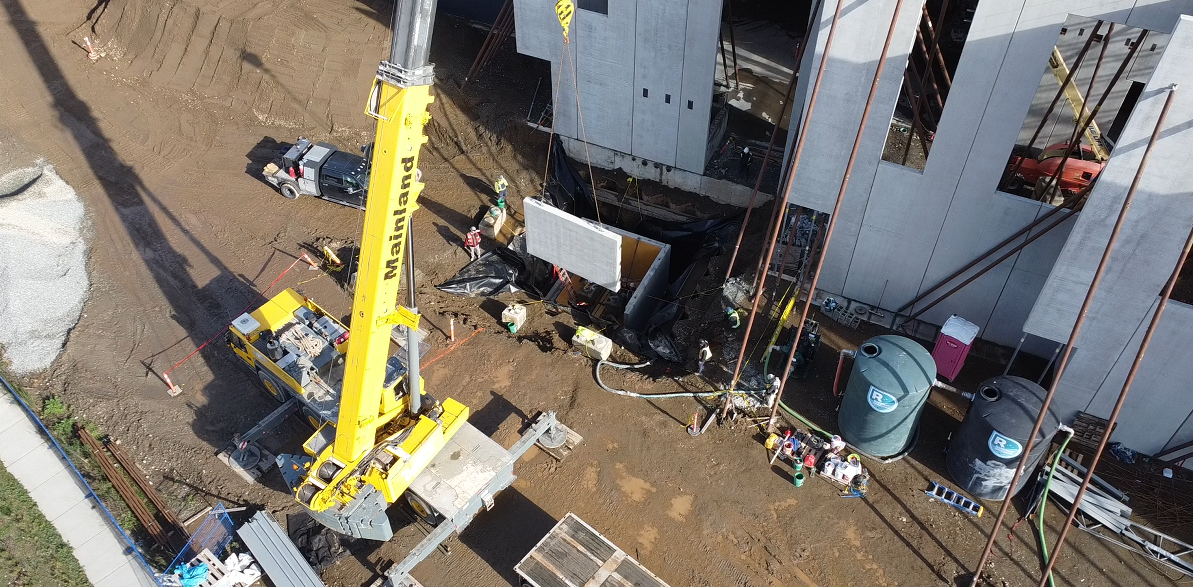 Aerial view of a building construction site with workers and a yellow crane lifting a concrete wall panel, surrounded by dirt, construction materials, and equipment.