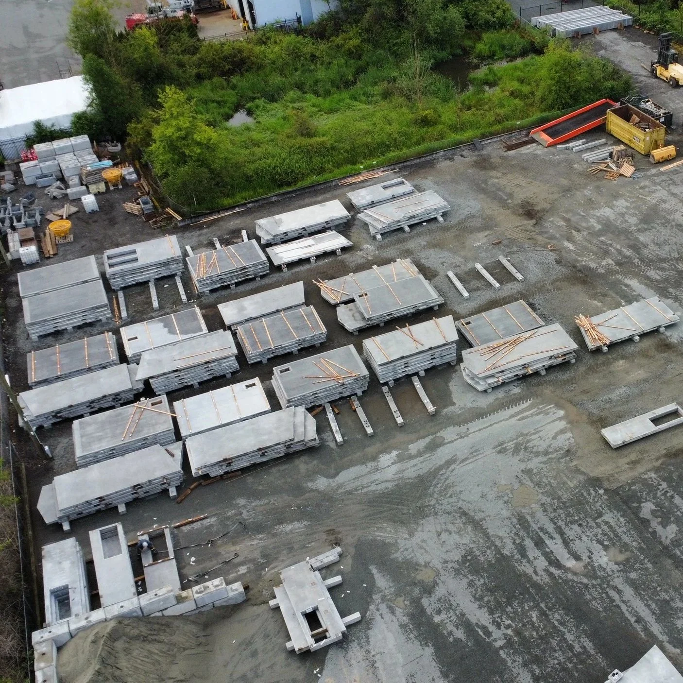 Aerial view of a precast concrete manufacturing yard with stacks of concrete blocks, metal beams, and construction materials, adjacent to a green, wooded area.