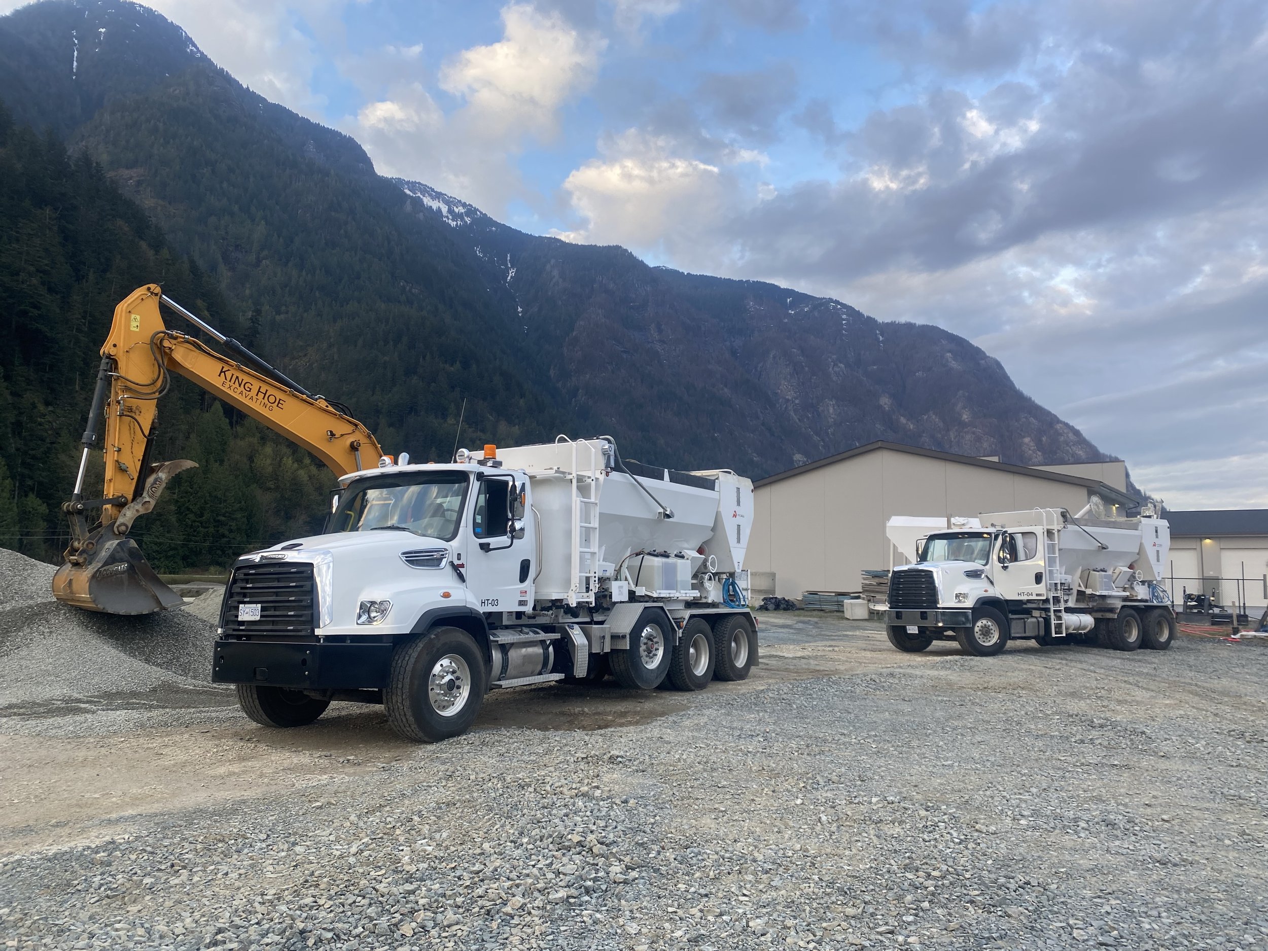 Construction equipment and trucks parked on a gravel lot with mountains and cloudy sky in the background.