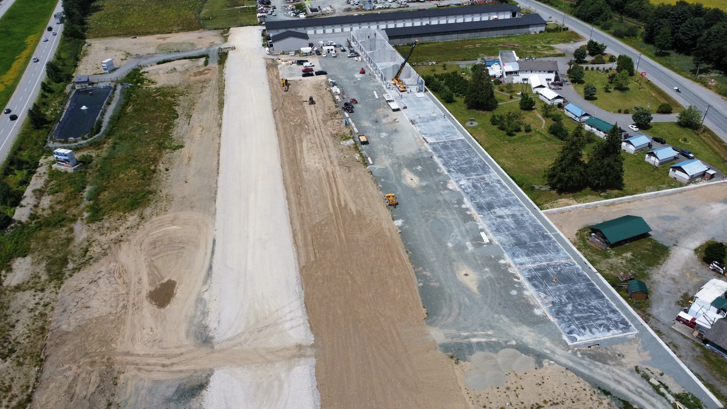 Aerial view of a construction site with partially paved parking lot, construction equipment, and vehicles. Surrounding area includes roads, green lawns, trees, small houses, and buildings.