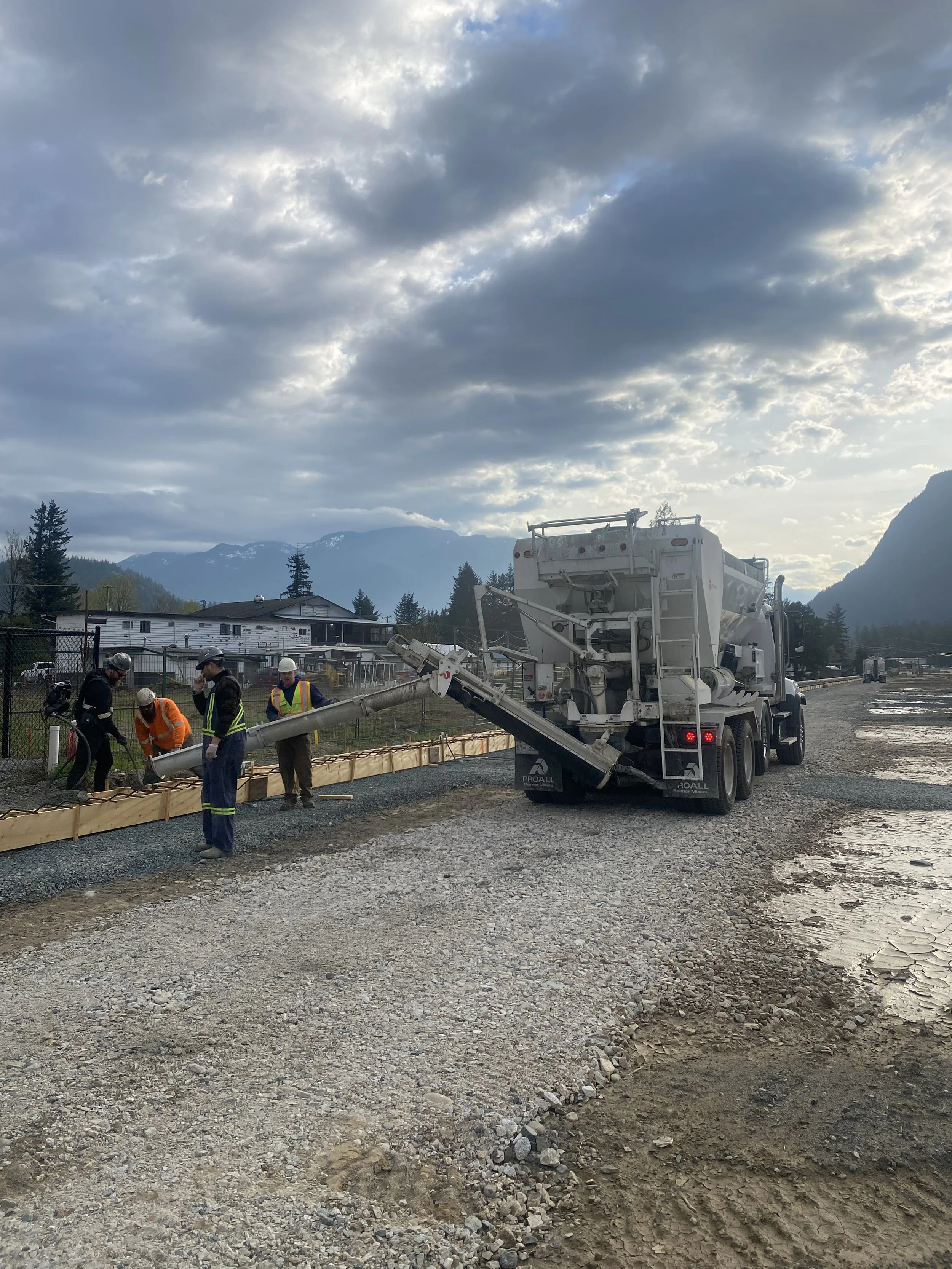 Construction workers are working on a project involving a concrete mixer truck and a wooden form for pouring concrete. The scene is outdoors with mountainous terrain and clouds in the sky, and the photo is taken from ground level in Hope, BC.

