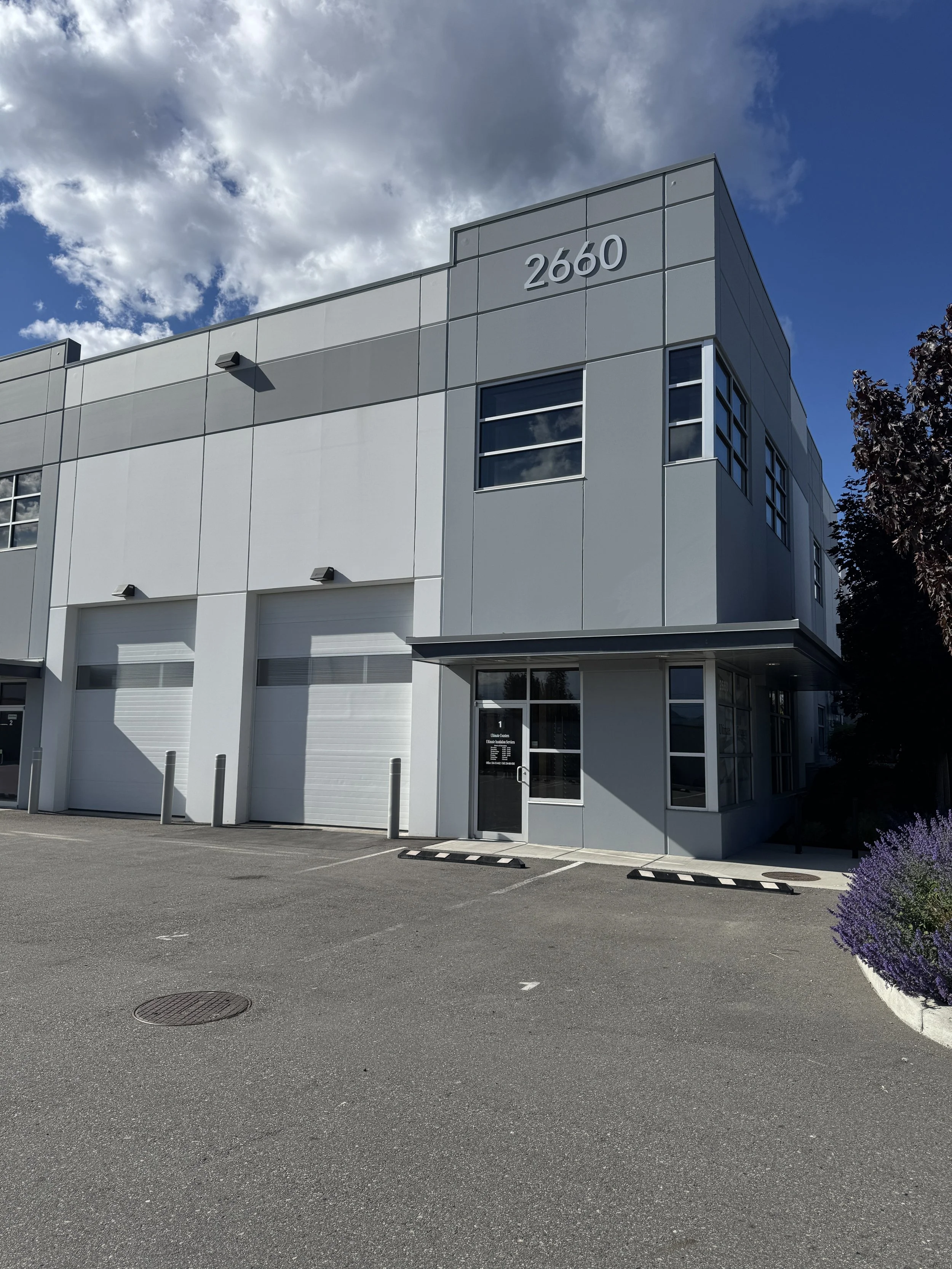 A modern, white commercial building with the number 2660 displayed on the corner, multiple garage doors, parking spaces in front, and a tree with purple flowers on the right, under a partly cloudy sky. The building is made of tilt-up concrete panels.