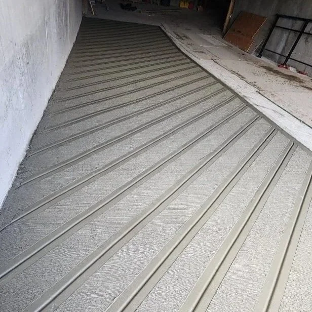 A newly installed textured concrete floor leading down to an apartment building's parking garage, adjacent to a white wall and a construction area with tools and unfinished surfaces.