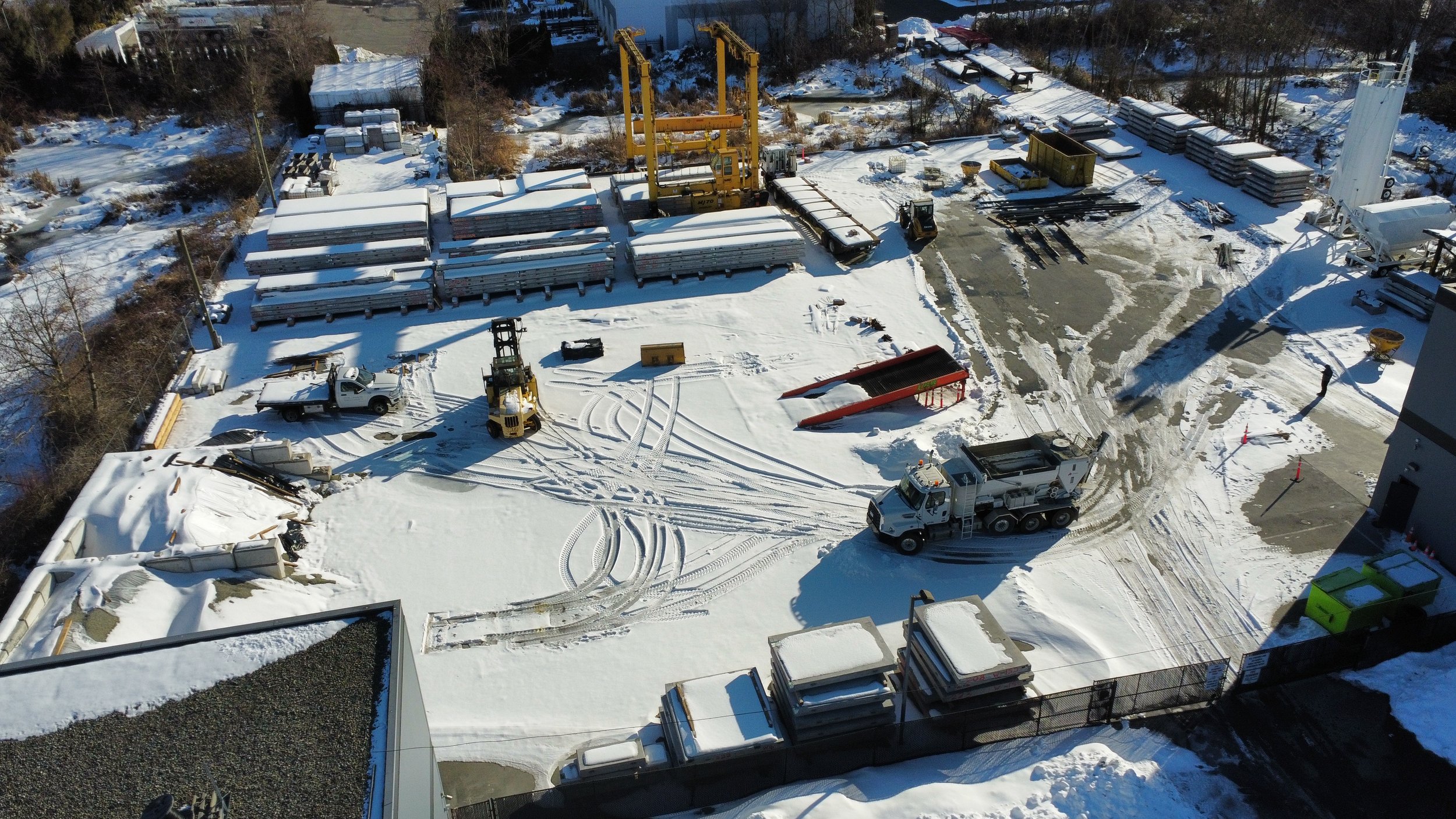 Baseline Concrete's Langley yard covered in snow with various construction equipment, vehicles, and materials, including a crane, forklifts, and stacks of building supplies, with tire tracks in the snow and a few workers present.