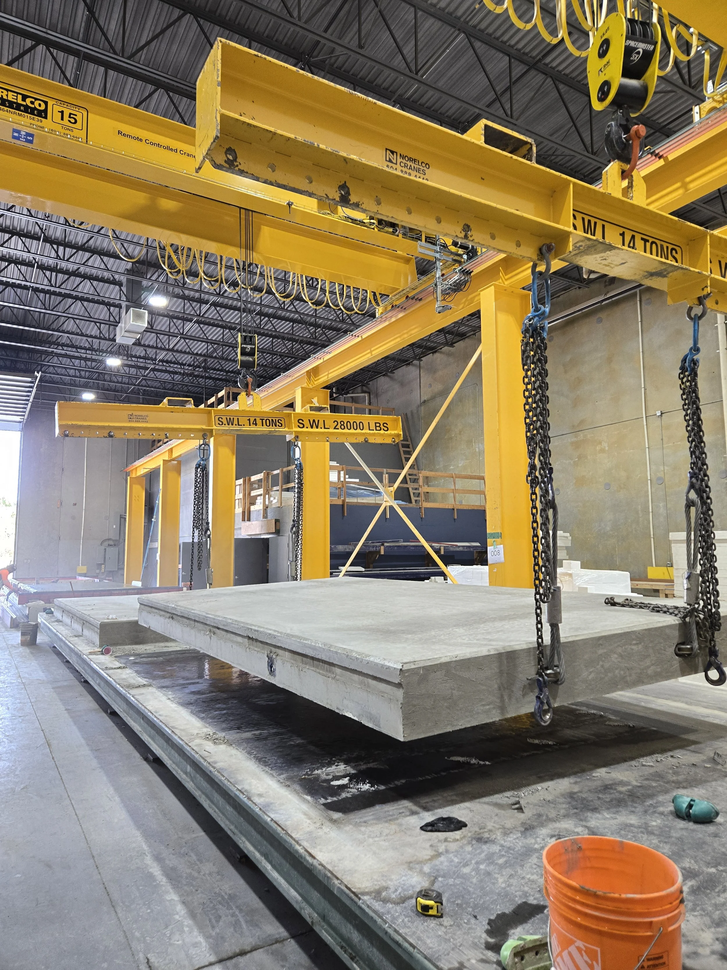 Large industrial crane in a warehouse lifting a precast concrete wall panel. The crane is painted yellow with weight specifications marked. The warehouse has high ceilings with visible metal framework and lights.