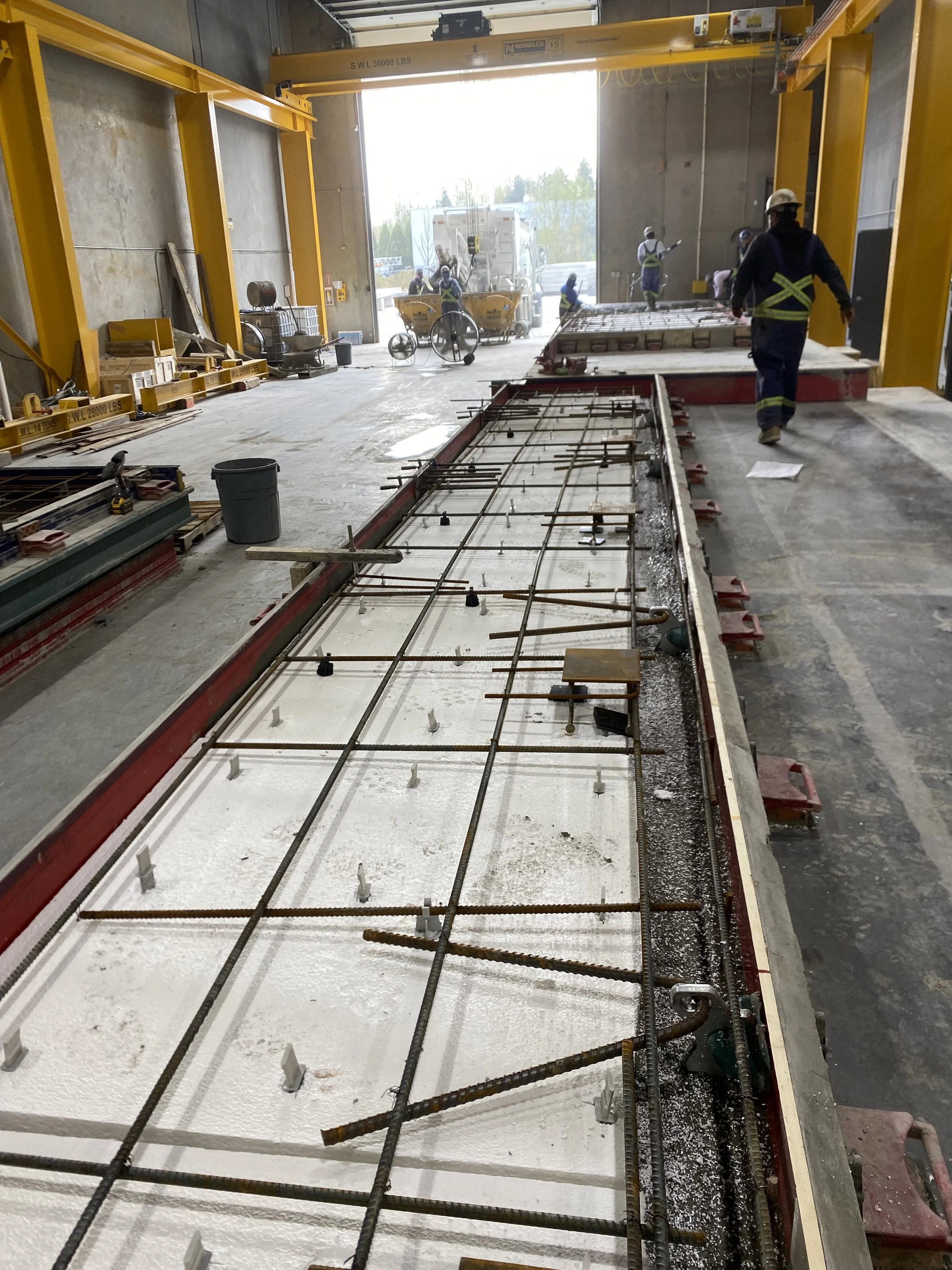 Construction workers are working inside a large industrial building, preparing a concrete slab with rebar reinforcement for pouring. A hoist is overhead, and various construction tools and materials are scattered around.