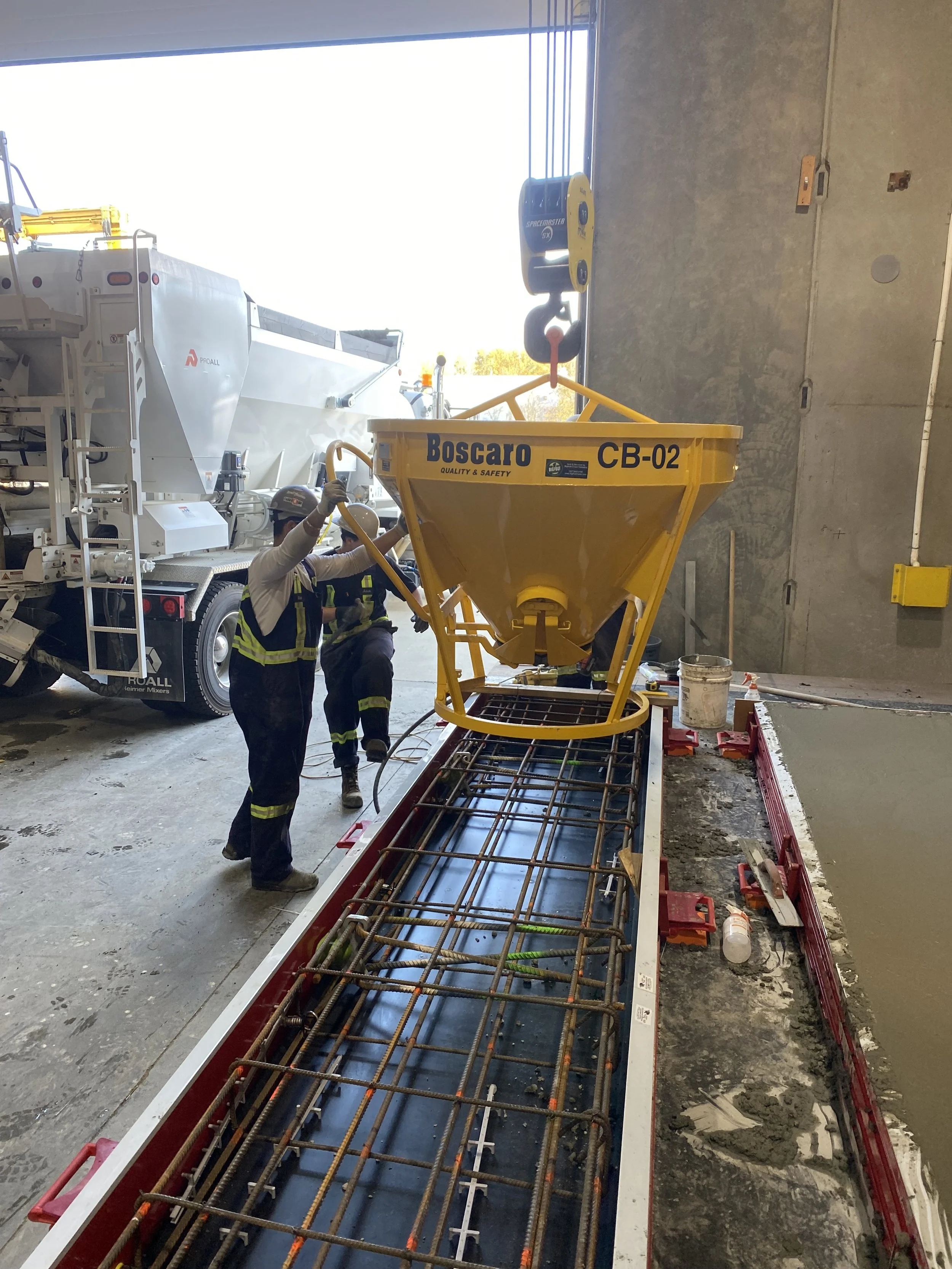 Workers in safety gear are pouring concrete into a wooden frame with rebar reinforcements.