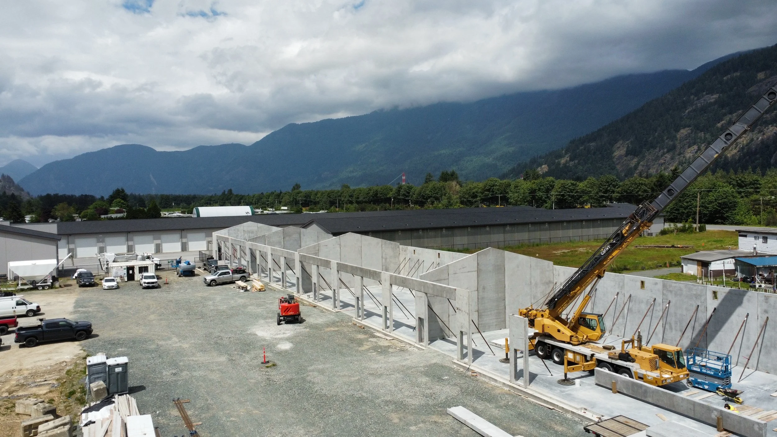 Construction site with vehicles, concrete panels, and a crane, set against green mountains and cloudy sky.