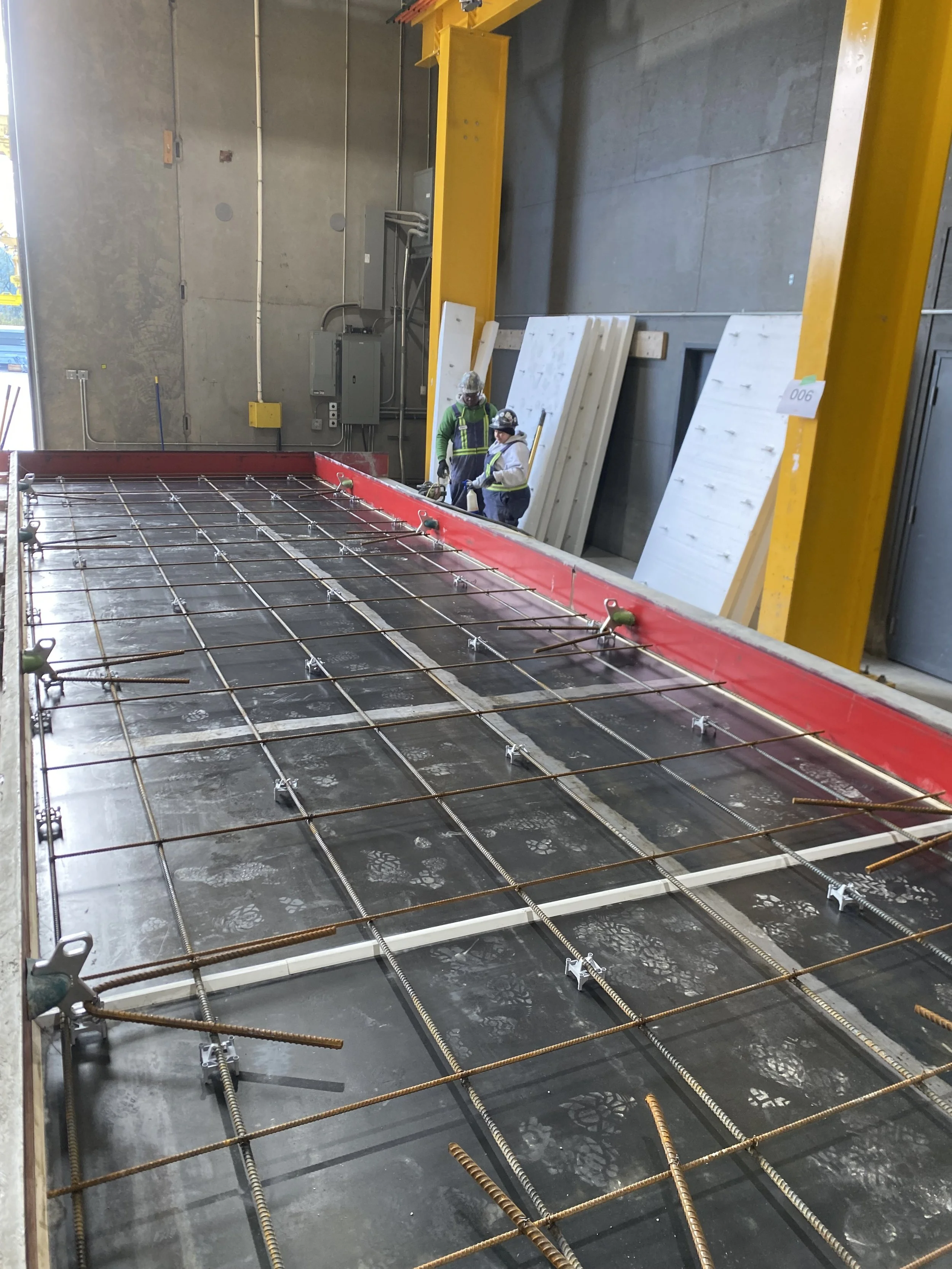 Two workers in safety gear and helmets working inside a concrete warehouse, with rebar and concrete preparations visible for a concrete pour, and large wooden boards behind them.