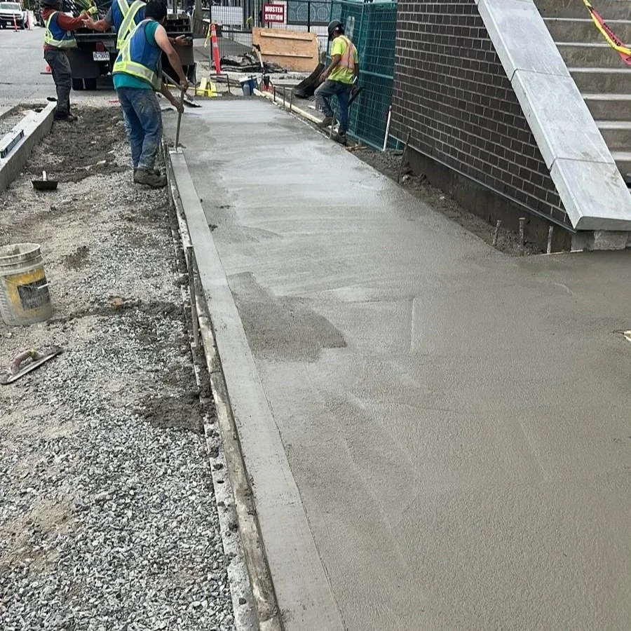 Workers in safety vests and helmets are smoothing freshly poured concrete for a sidewalk or pathway outside a building under construction in Downtown Langley, BC.