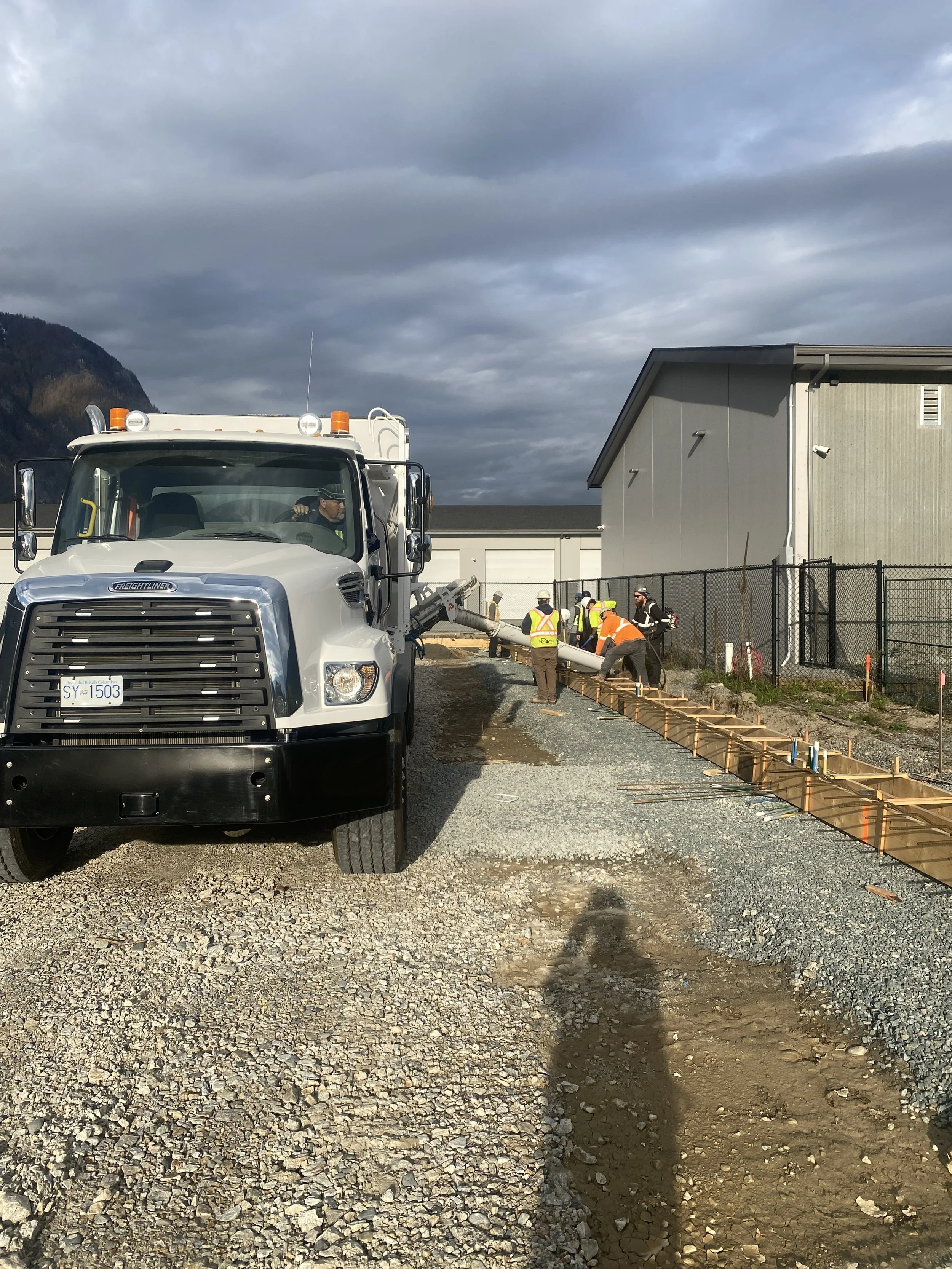 A photo of a Volumetric concrete truck at a building site with gravel and dirt ground, gray building, and dark cloudy sky in the background.