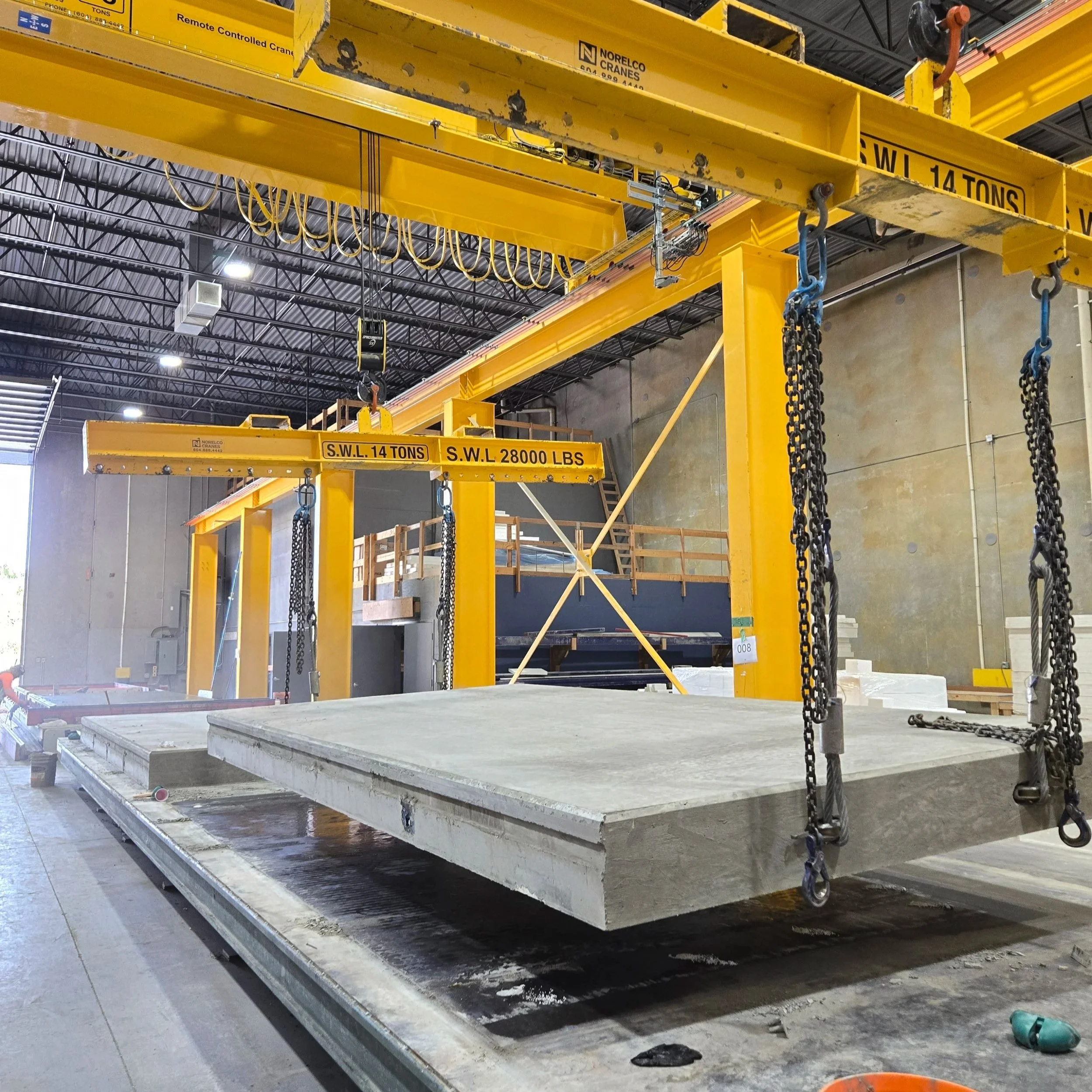 Large industrial crane in a warehouse lifting a precast concrete wall panel. The crane is painted yellow with weight specifications marked. The warehouse has high ceilings with visible metal framework and lights.