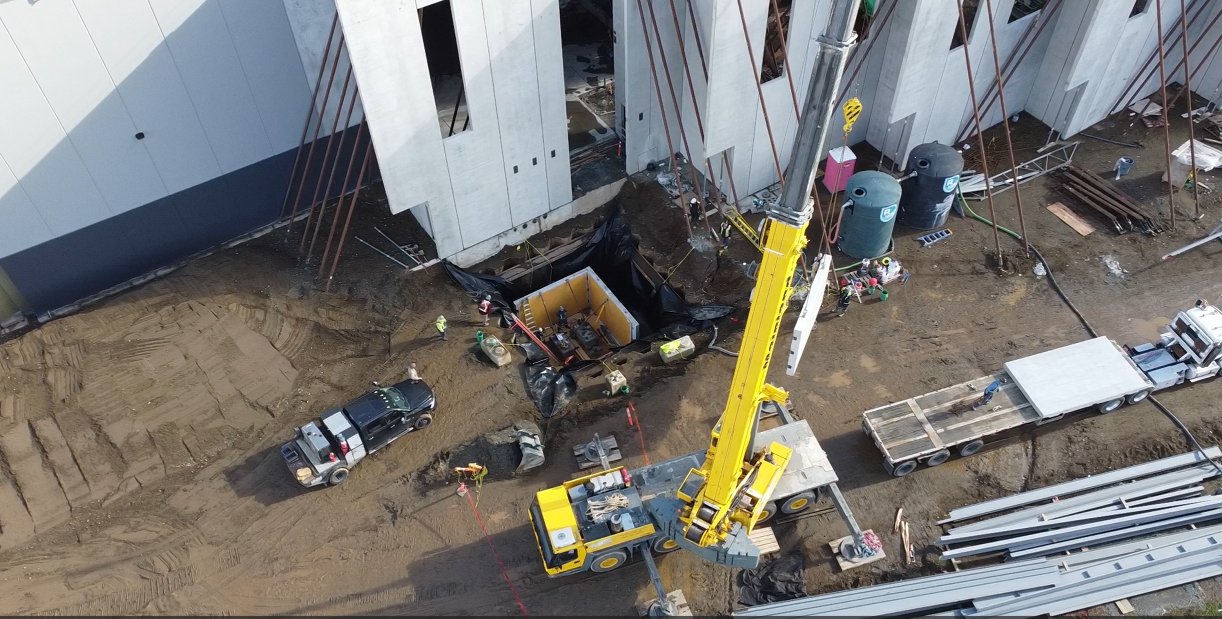 Construction site with a large yellow crane, workers, construction materials, and a partially built wastewater chamber with gray concrete walls and exposed framing.