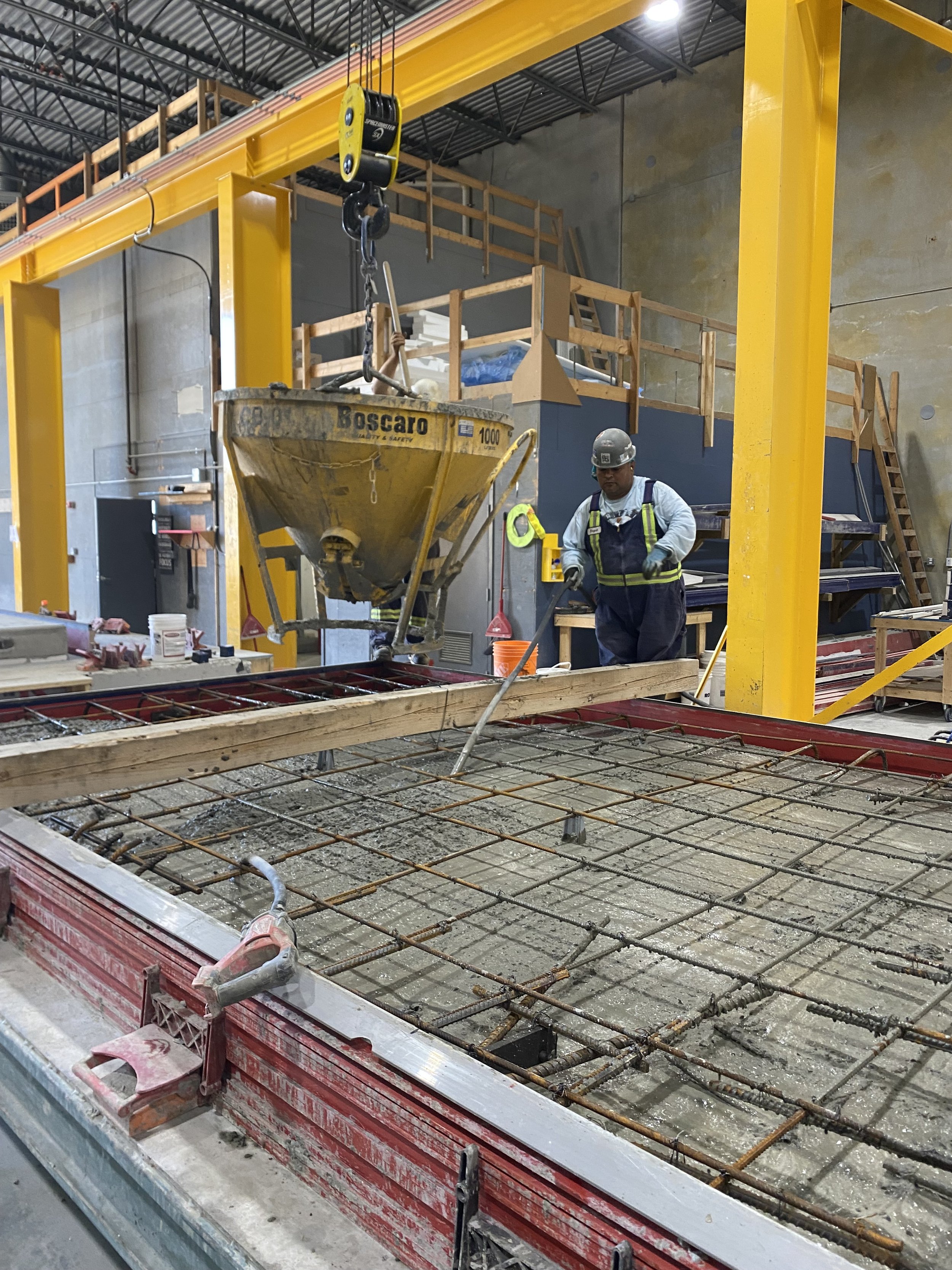 Construction worker in a gray uniform and helmet working on a concrete slab with rebar inside a warehouse building with exposed concrete walls.