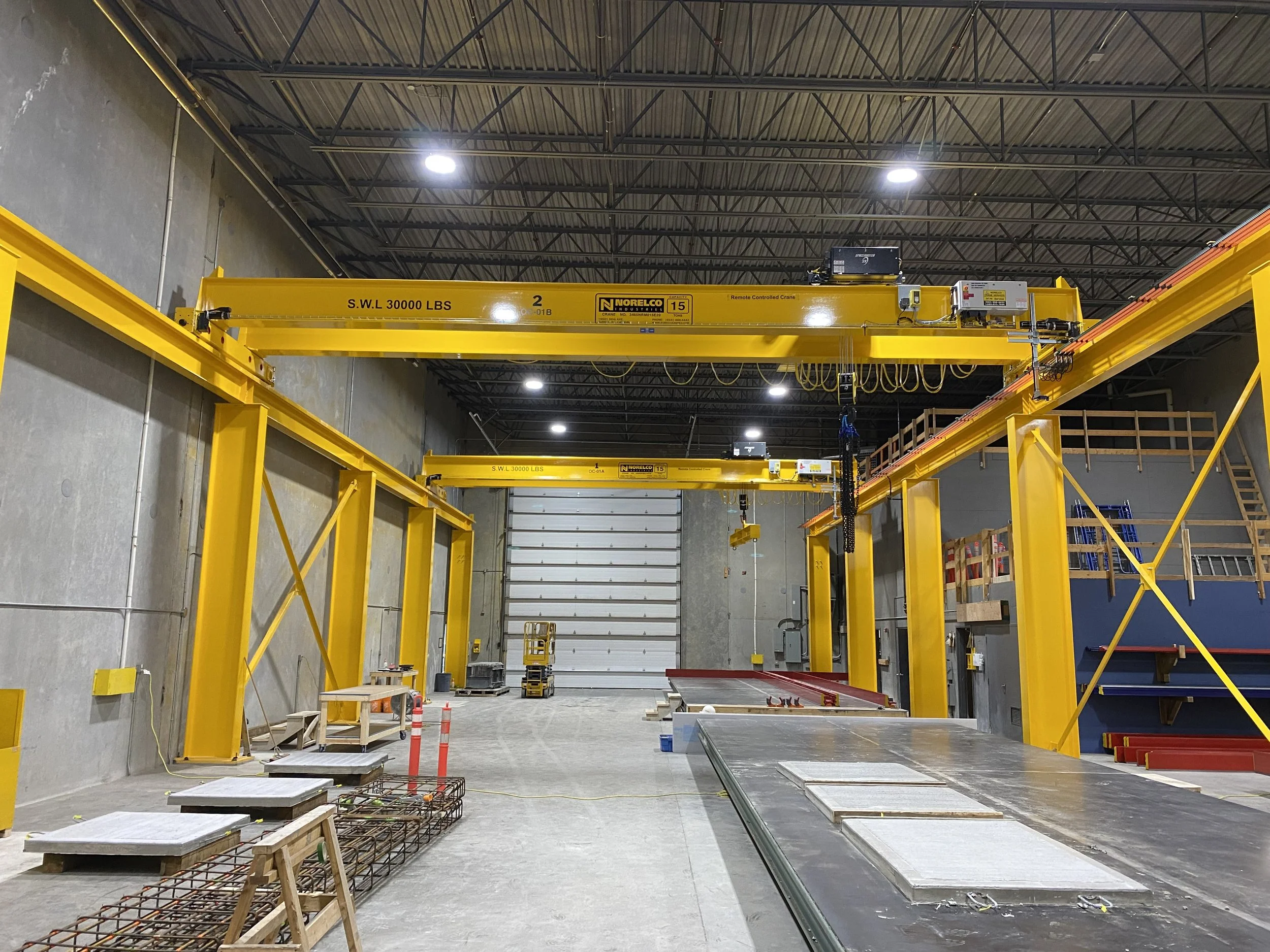 An industrial concrete warehouse at A-5350 272 St, Langley, BC,  with yellow overhead cranes, construction materials, and tools, set against a high ceiling with metal beams and ceiling lights.