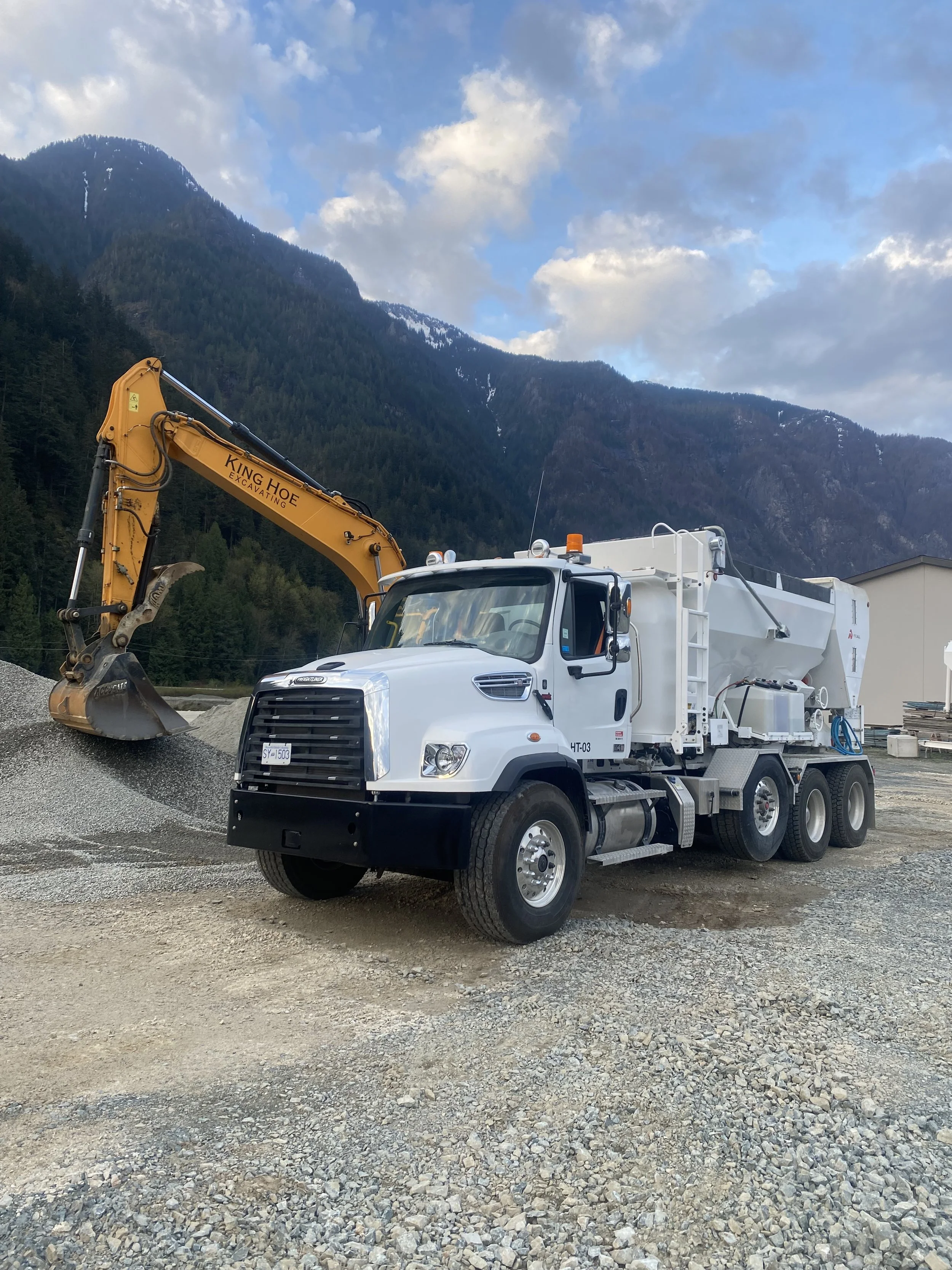 A volumetric concrete truck parked on a construction site with a white truck and a yellow excavator against a mountain landscape with cloudy sky.