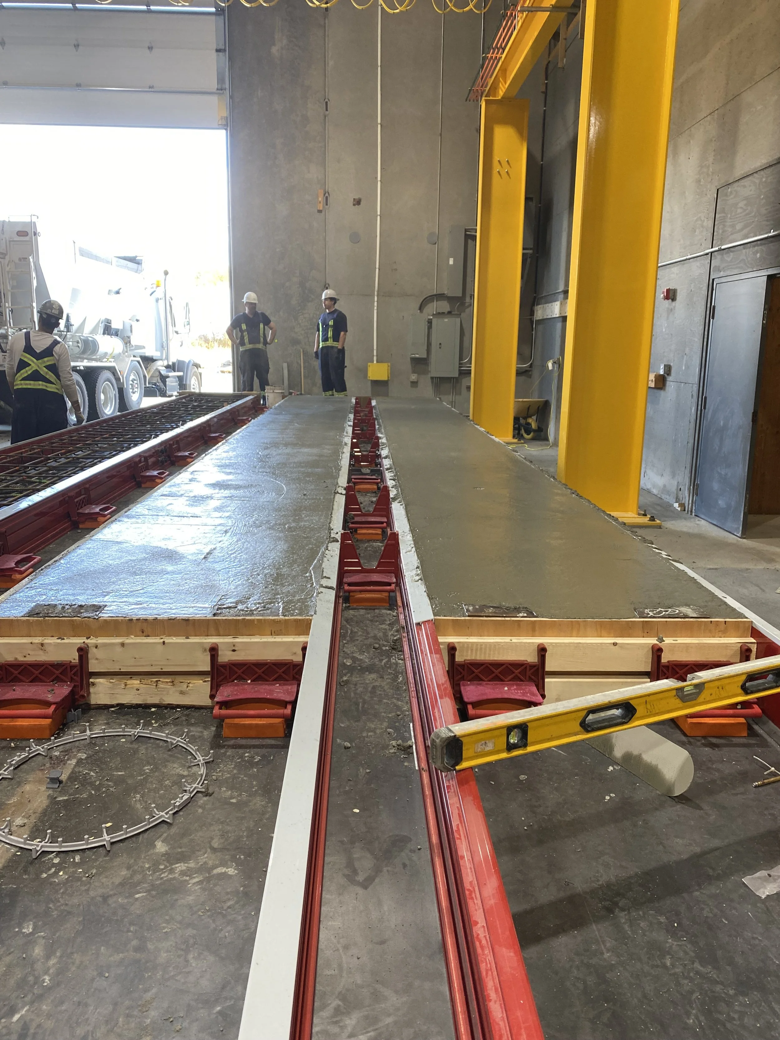 Construction workers wearing safety helmets and vests standing inside a large industrial or warehouse space, with a concrete floor, yellow safety barriers, and a large door opening to the outside. There is construction equipment and materials, includ