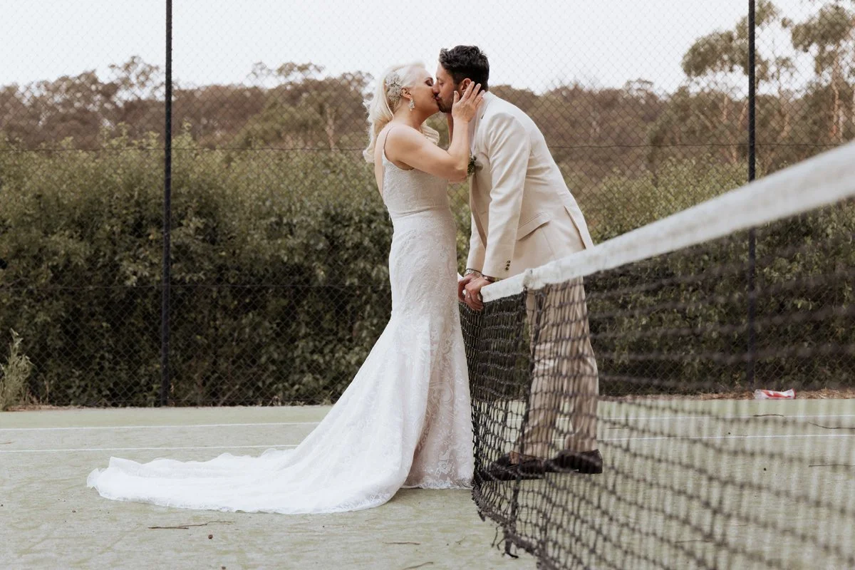 Editorial wedding photographer Melbourne - A bride and groom kissing on a tennis court, holding hands, with a tennis net in the foreground and trees in the background.