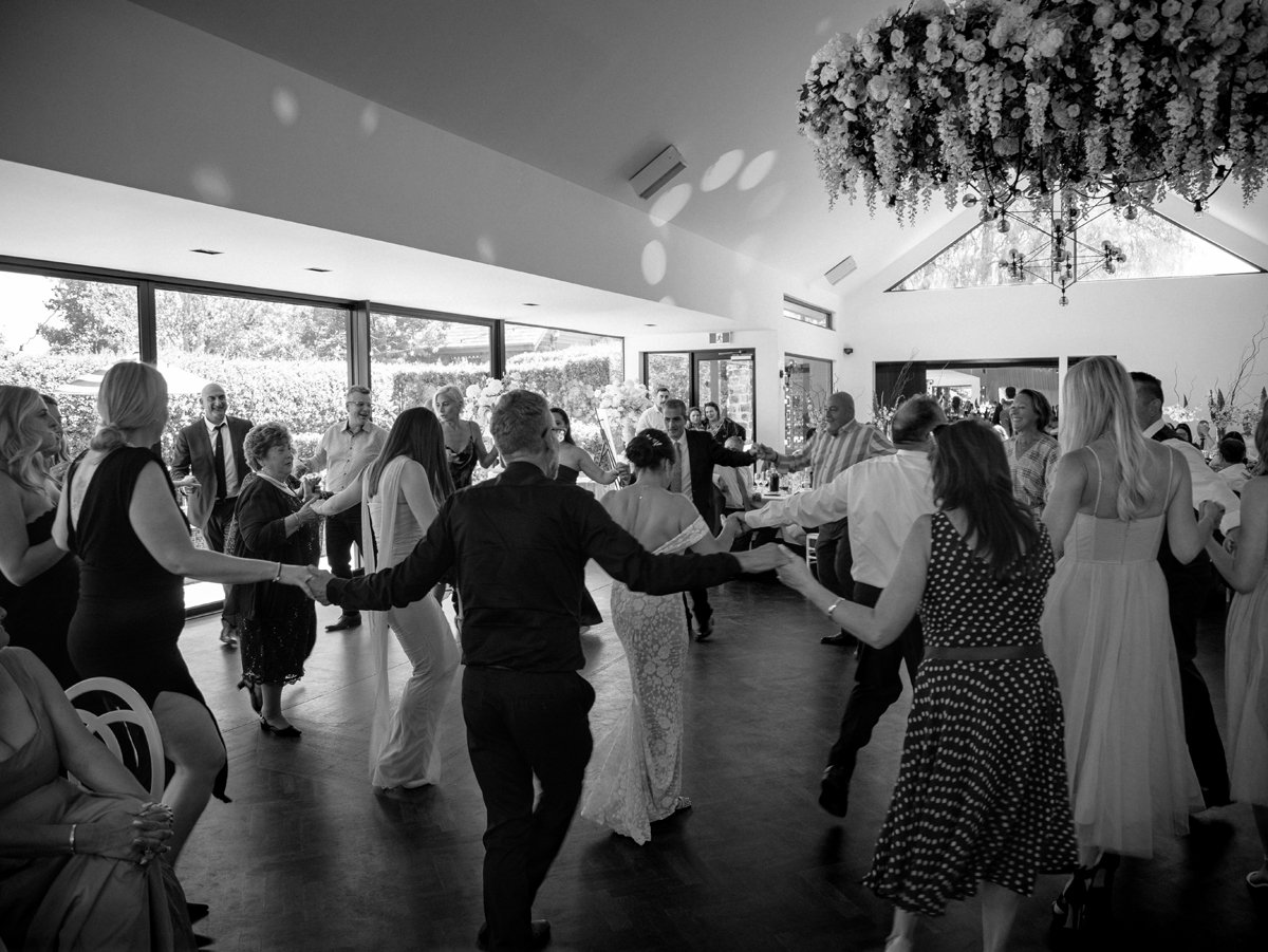 Editorial wedding photographer Melbourne - People dancing in a circle inside a decorated wedding reception venue with large windows and natural light.