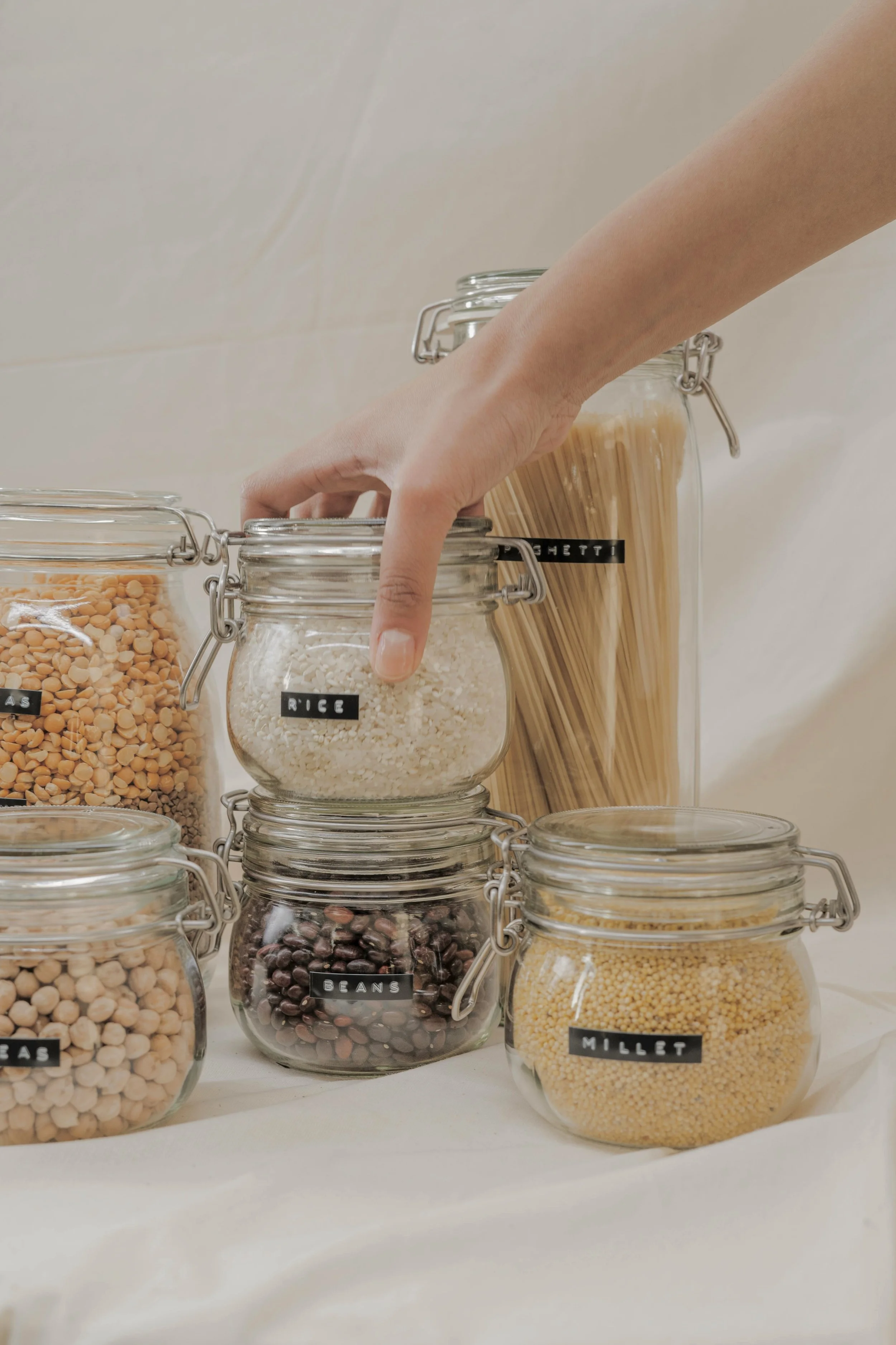 A hand reaching for a jar labeled rice among other jars of beans, millet, and uncooked pasta.